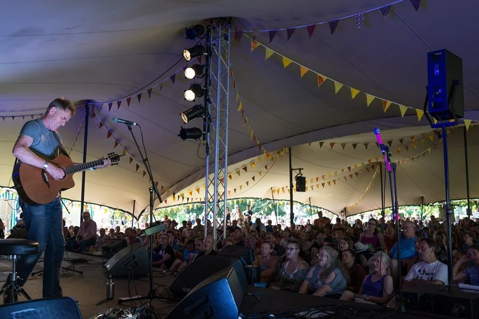 A musician playing a guitar on a stage in a large stretch tent with an audience watching, decorated with colourful triangular banners.