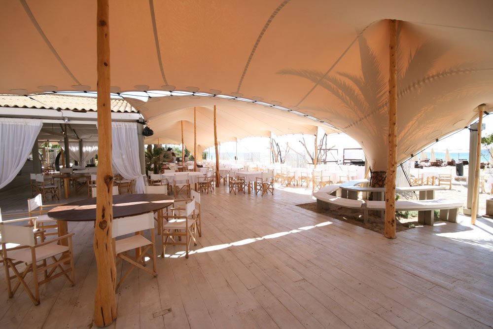 Open-air restaurant or event space with wooden floors, white tables, and chairs, under a large beige fabric canopy supported by wooden poles, overlooking a beach or waterfront area.