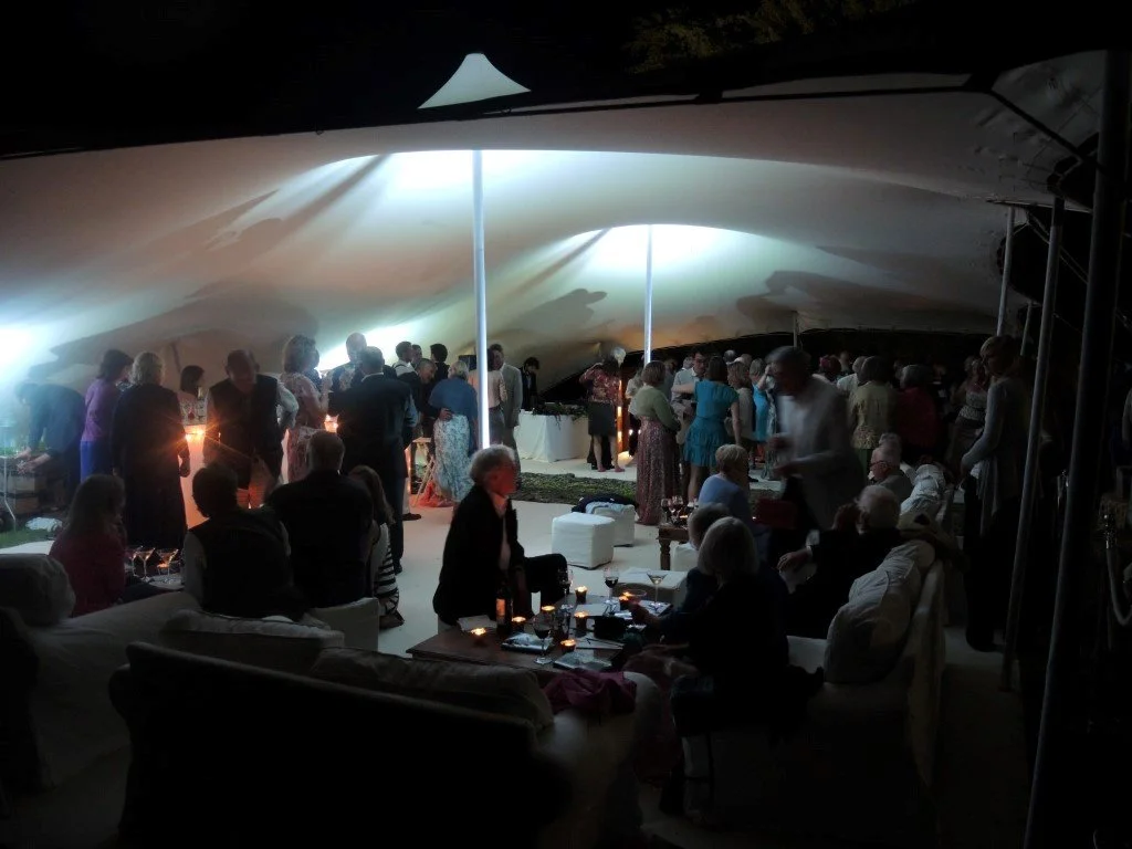 People gathered at a social event inside a large stretch tent with a modern, flowing ceiling design, some seated and others mingling around tables with candles and drinks.