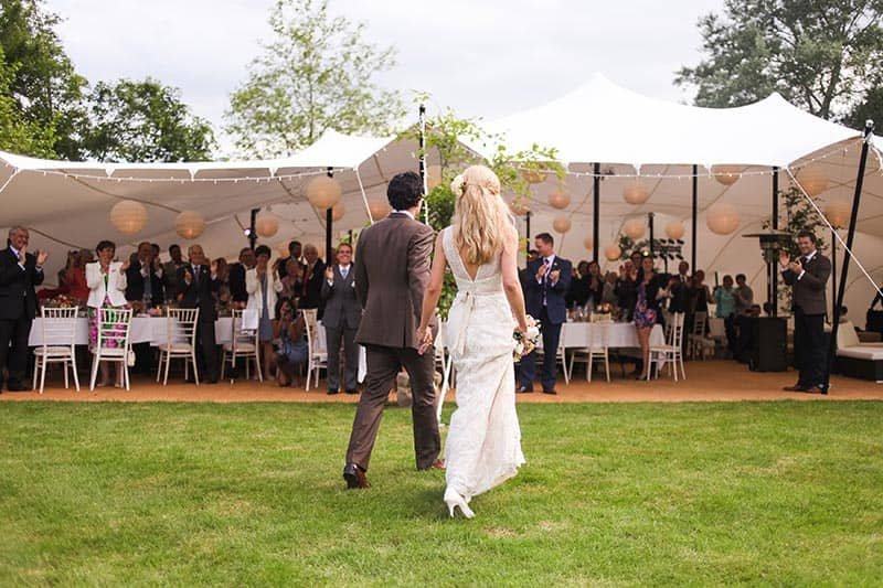 Couple walking towards a decorated outdoor wedding reception tent with guests inside, under a white canopy with hanging paper lanterns.