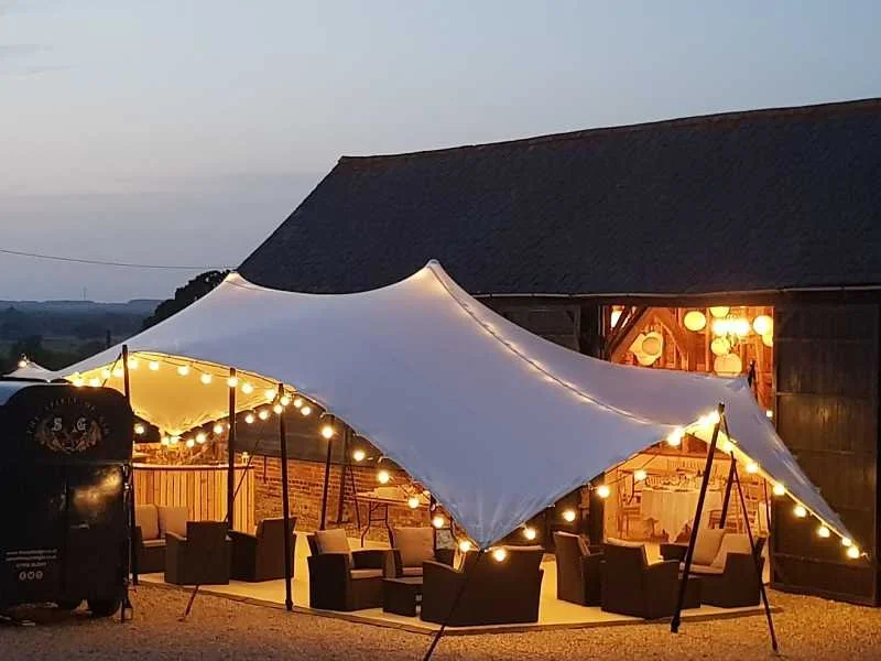 Outdoor event space with a white tent decorated with string lights, seating area with chairs and tables, set against a rustic barn at dusk.
