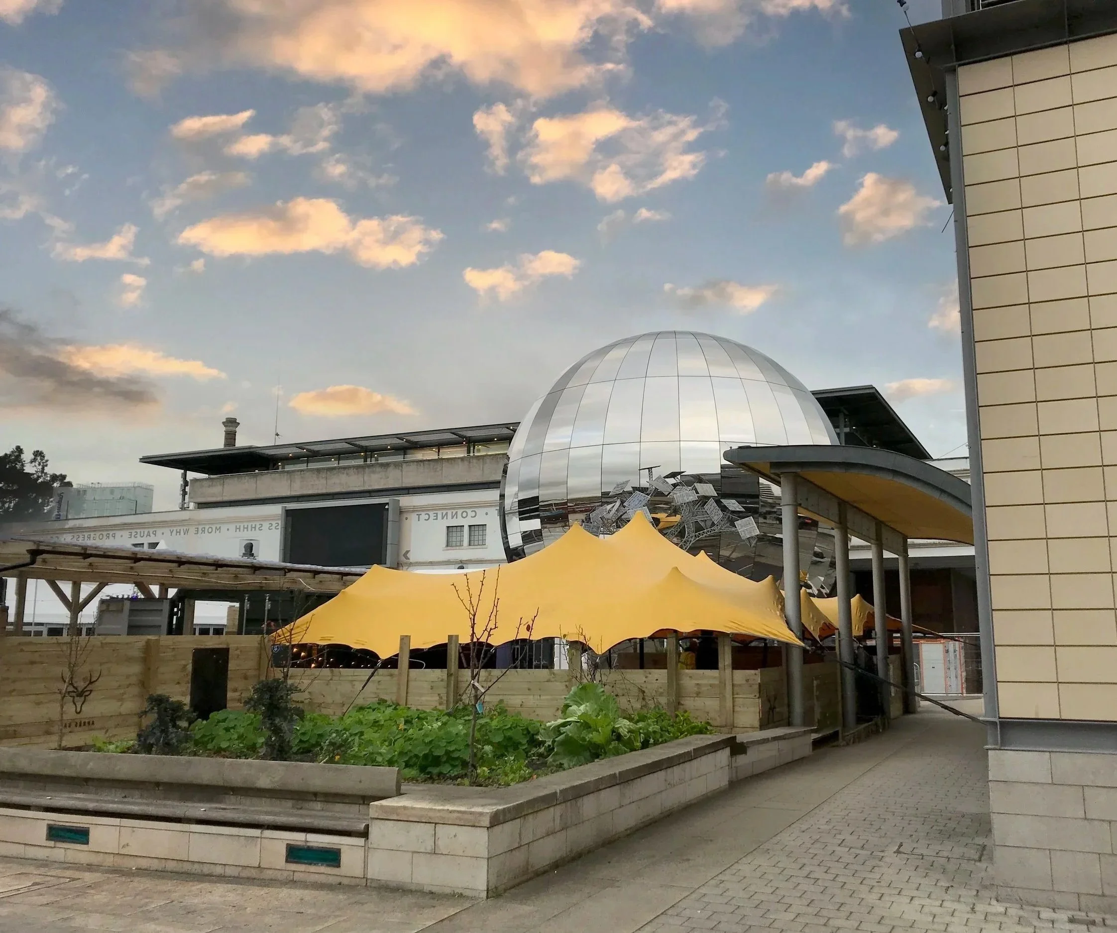 Urban scene with a modern building featuring a shiny, reflective dome and a yellow stretch tent in a small garden area.
