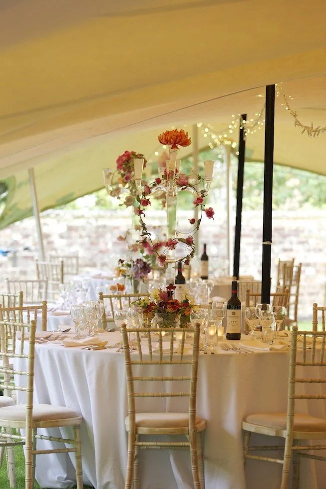 Elegant stretch tent with round table, white tablecloth, floral centrepiece, and gold chairs, under a tent with string lights.