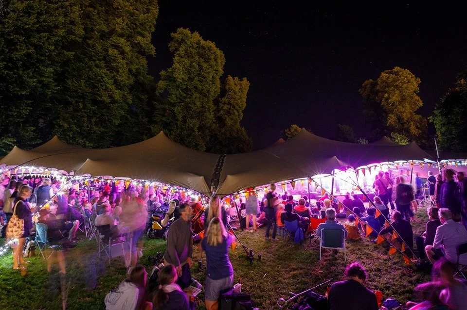 Night outdoor event with many people under a large canopy decorated with string lights, surrounded by trees.