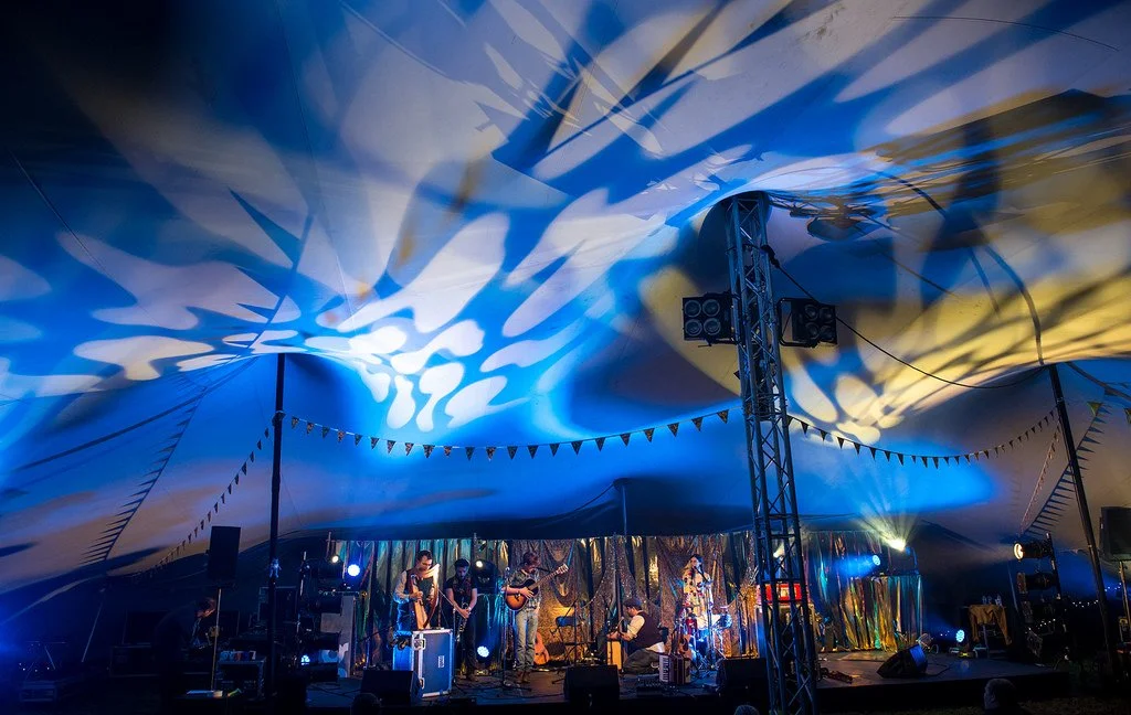 Music band performing on stage inside a decorated stretch tent with blue and yellow lighting.