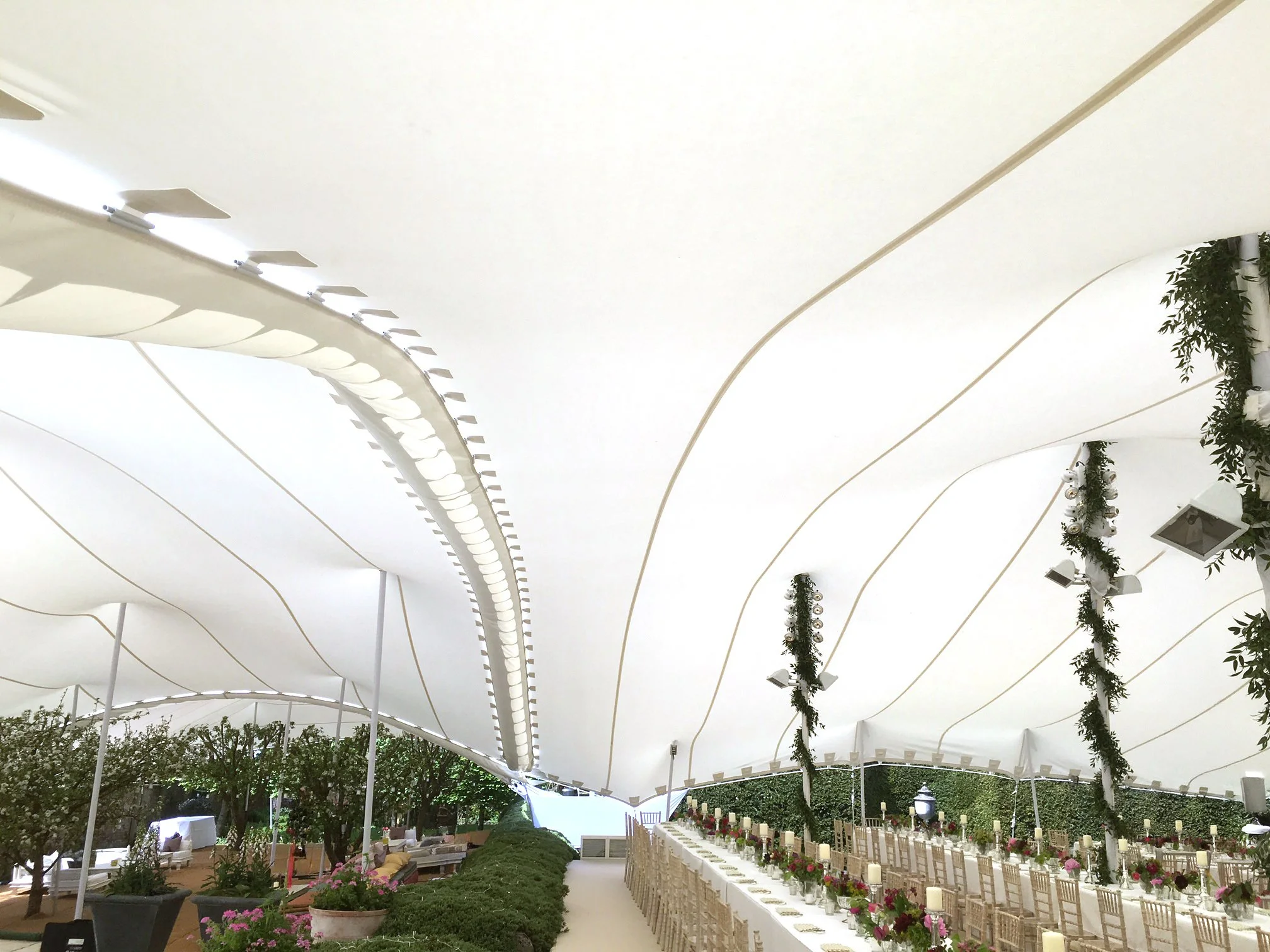 Indoor event space decorated with long banquet tables, white chairs, floral centerpieces, candles, and greenery, under a large, white, stretch tent with hanging, garland decorations.