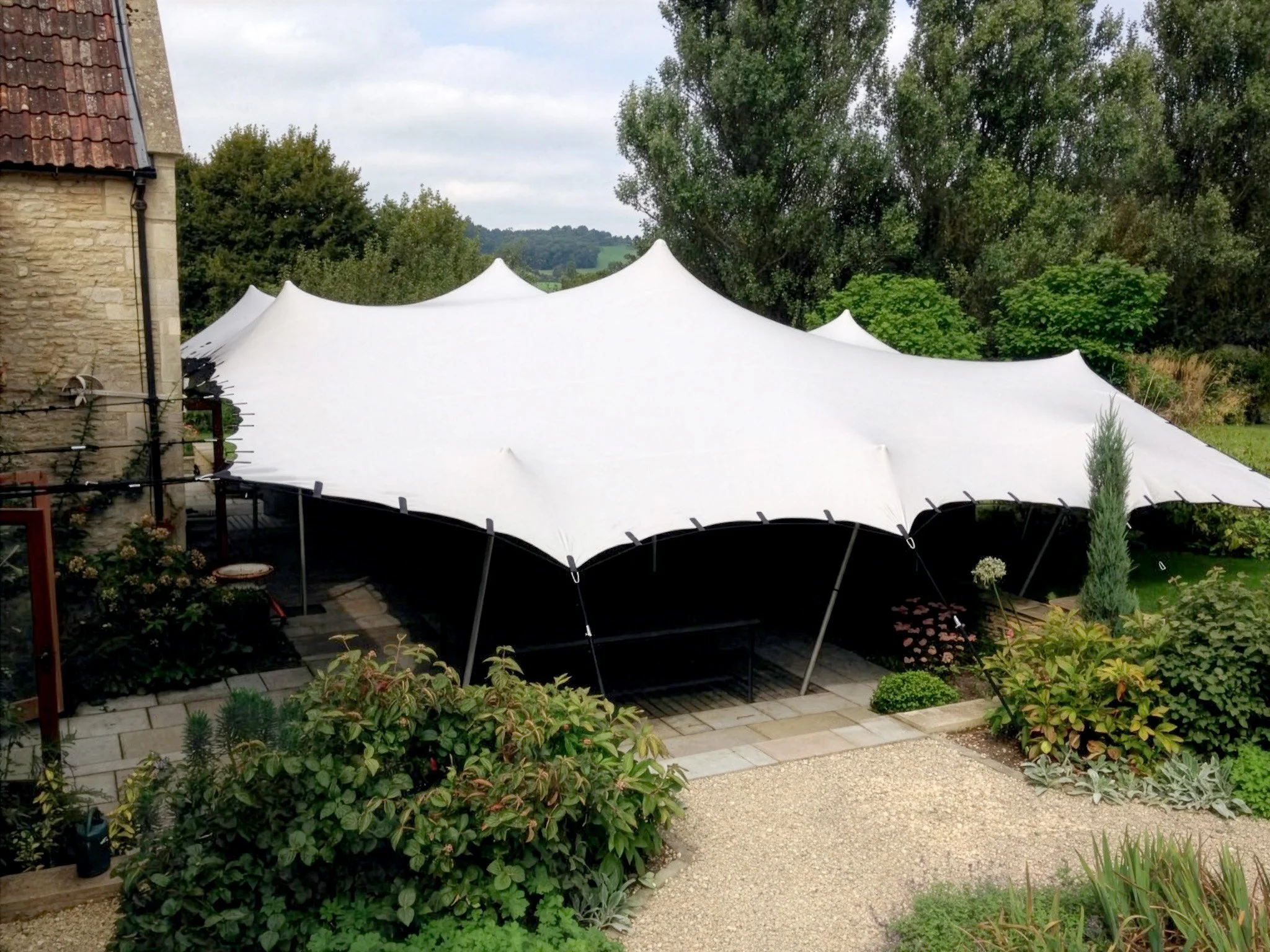 A large outdoor white stretch tent set up in a garden with greenery and trees in the background.