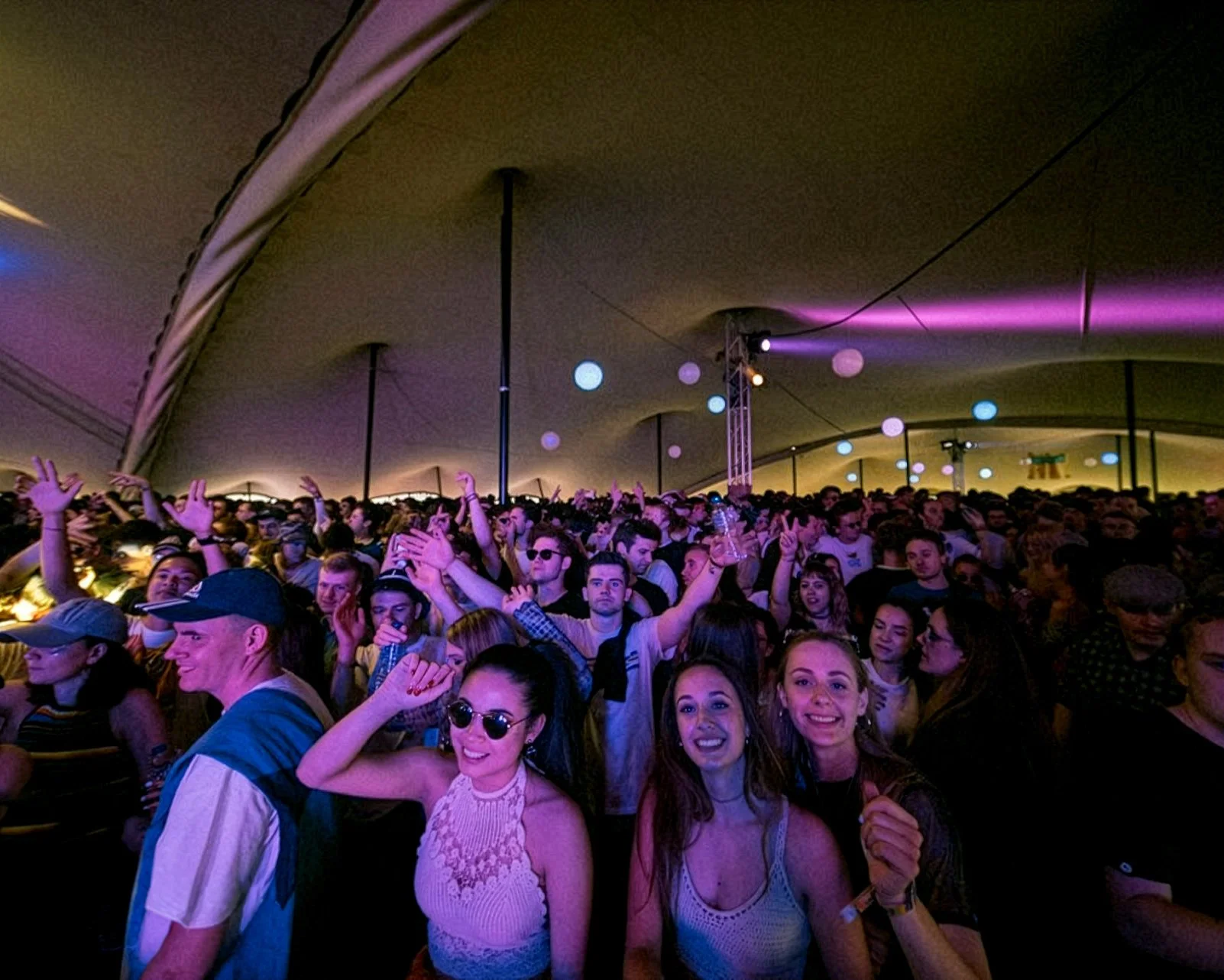 Crowd of people enjoying a concert or festival inside a large stretch tent with colourful lighting and hanging paper lanterns.