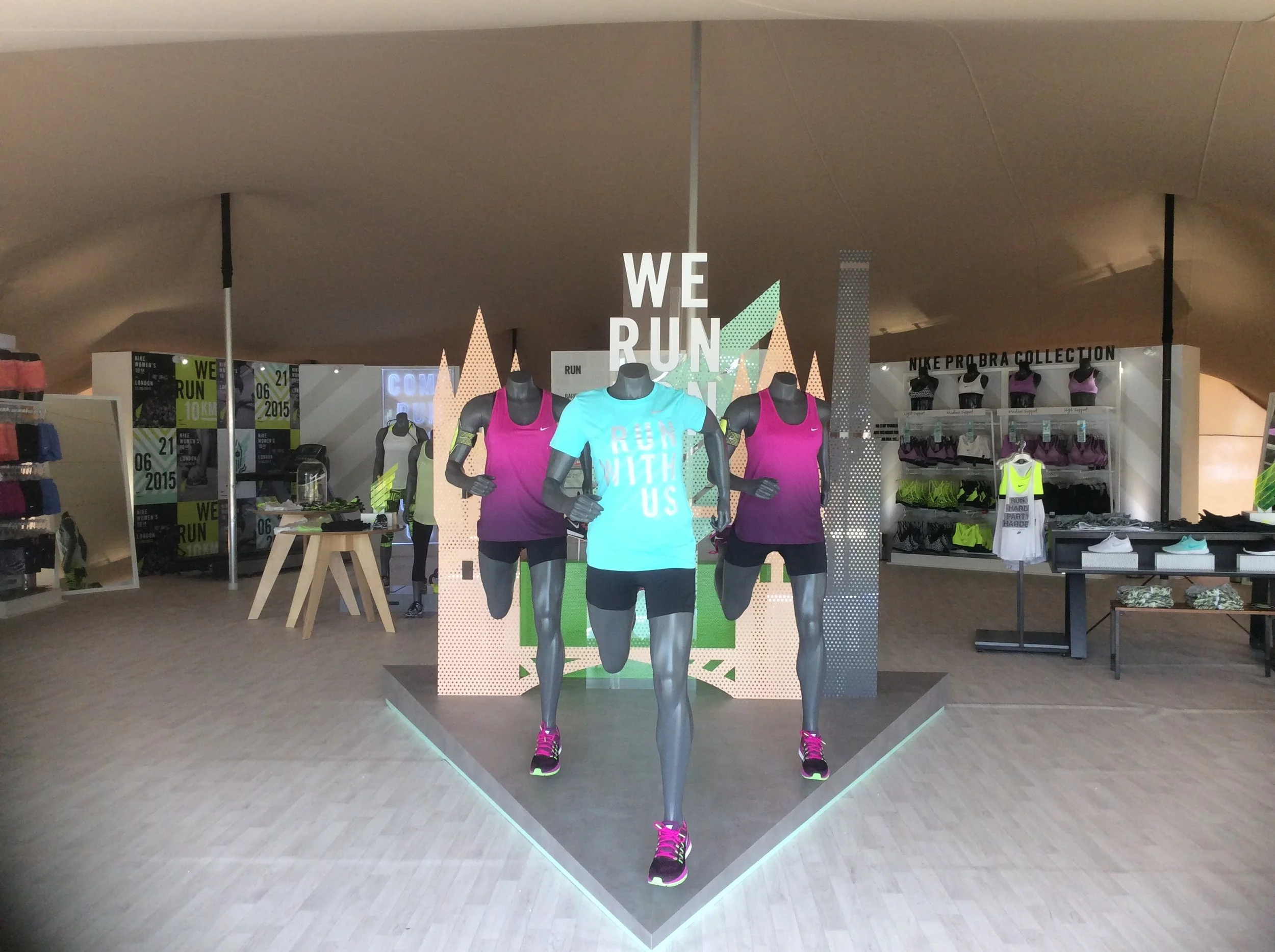 Display of sportswear mannequins at a stretch tent, with a prominent sign reading 'WE RUN' and colorful athletic clothing, including pink, purple, and neon green running outfits, arranged on mannequins for promotion.