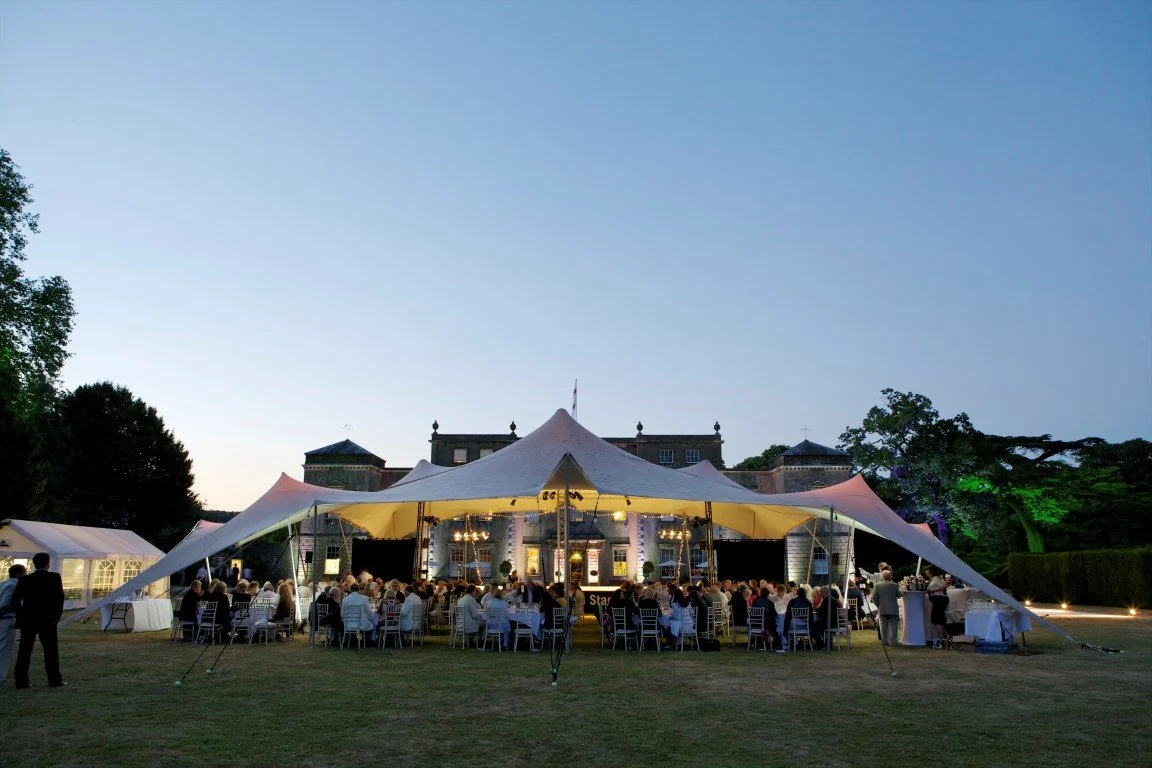 A large outdoor wedding reception under a white stretch tent with tables and chairs, set on the lawn of a grand estate at dusk.