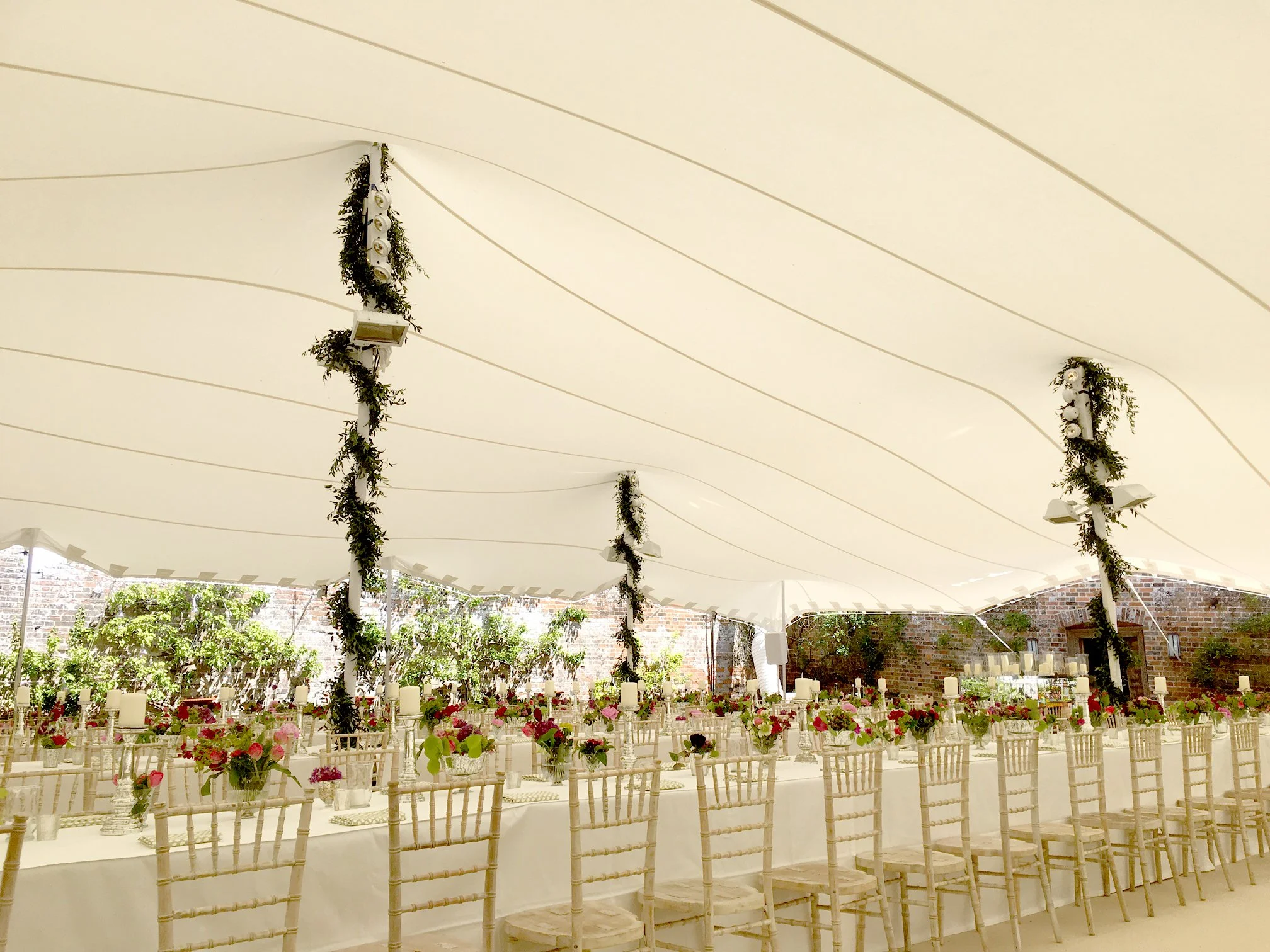 Interior of a large wedding tent decorated with flowers, candles, and draped fabric, with folding chairs arranged around tables for guests.