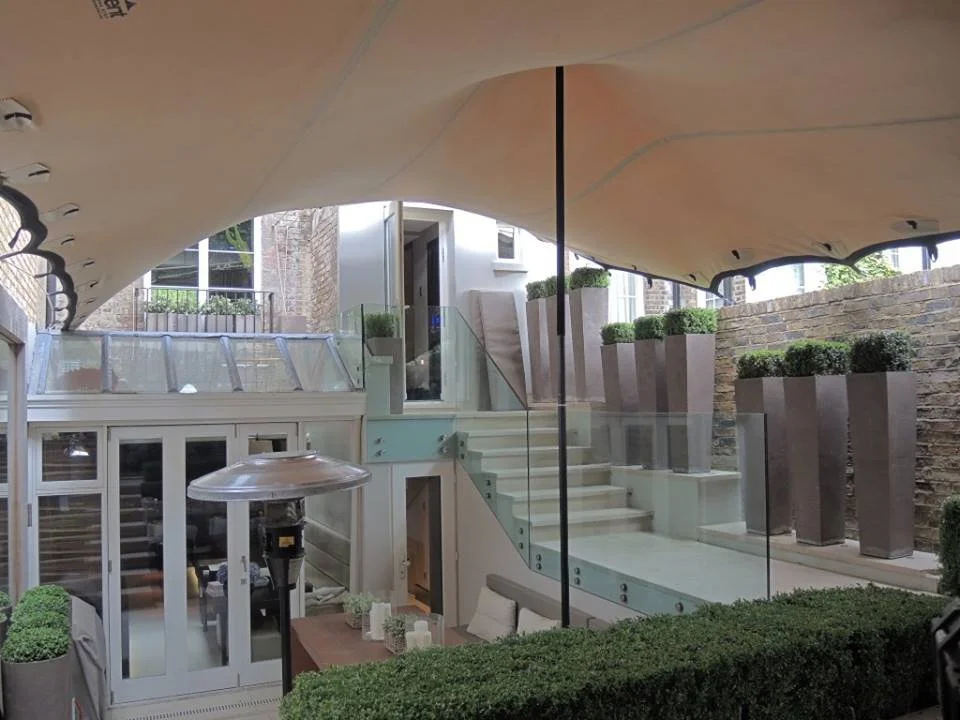 A modern multi-level outdoor patio with stairs, glass railings, large planters with green bushes, underneath a stretch tent.