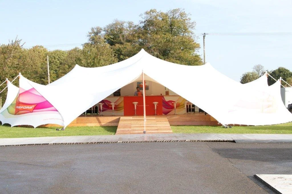 Large white stretch tent with colourful interior, set up on a grassy area, with a wooden stage and bar stools inside, and trees in the background.