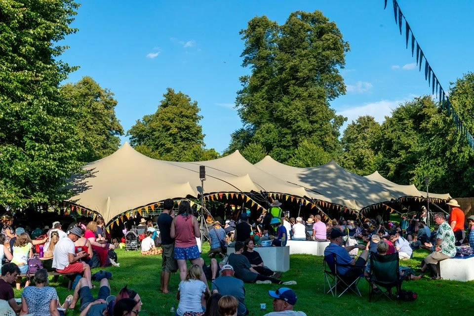 A lively outdoor festival with large stretch tents, people sitting on grass and chairs, trees, and a clear blue sky.