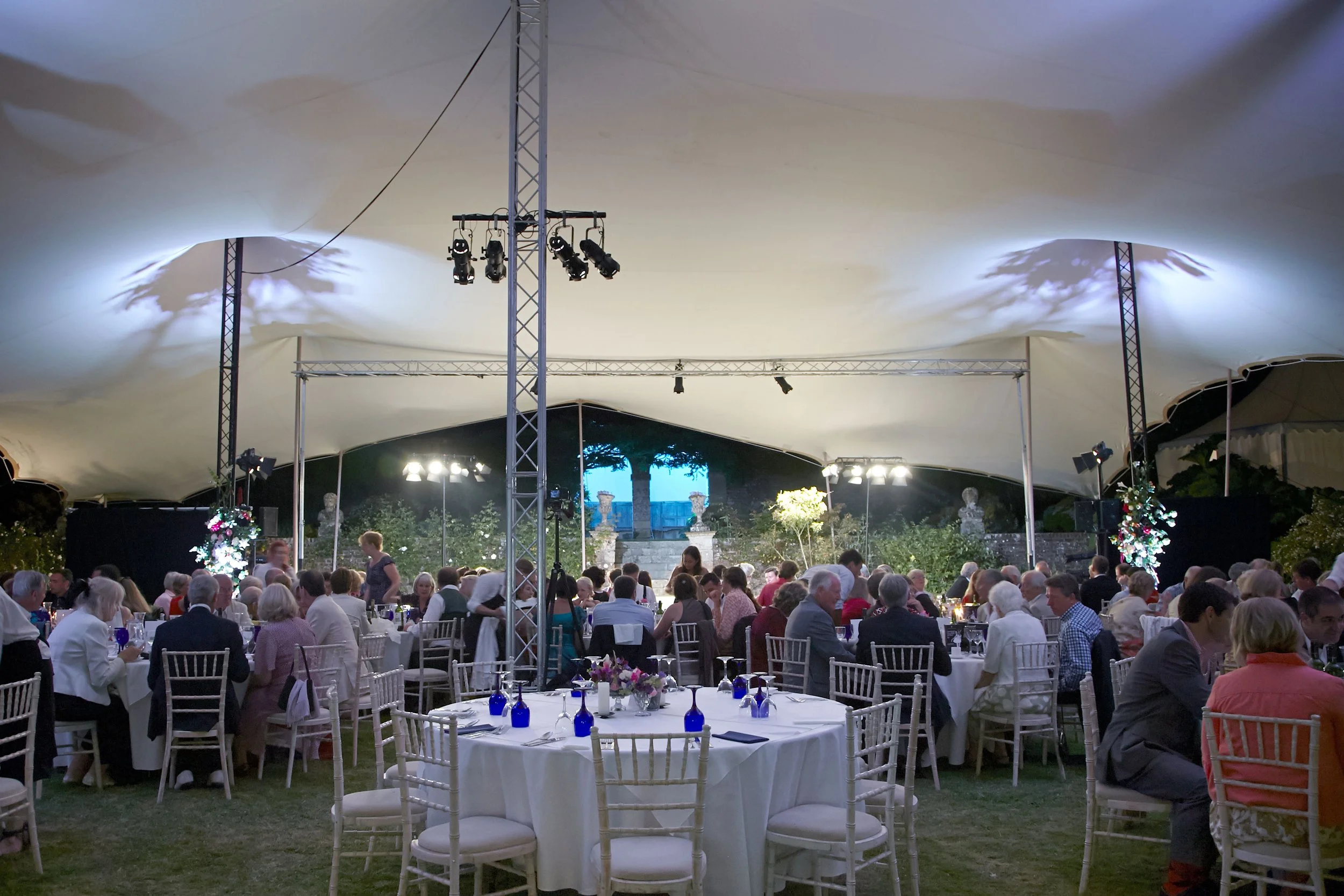 Guests seated at round tables under a large tent at an elegant outdoor event, with a stage and lighting at the front.