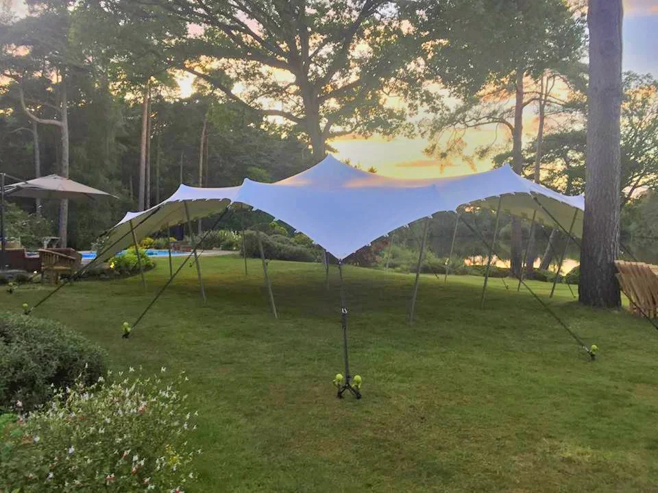 Large white stretch tent with multiple supporting poles set up on a grassy lawn during sunset, with trees and a lake or pond in the background.