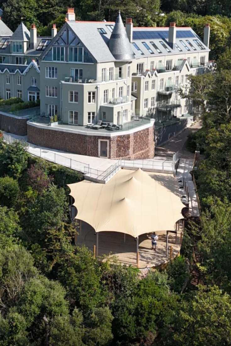 A large, light-colored multi-story building with steep roofs, turrets, and balconies, situated on a hillside surrounded by trees. In front of the building, there is a circular wooden deck with a large beige canopy, providing shade for a person standing underneath.