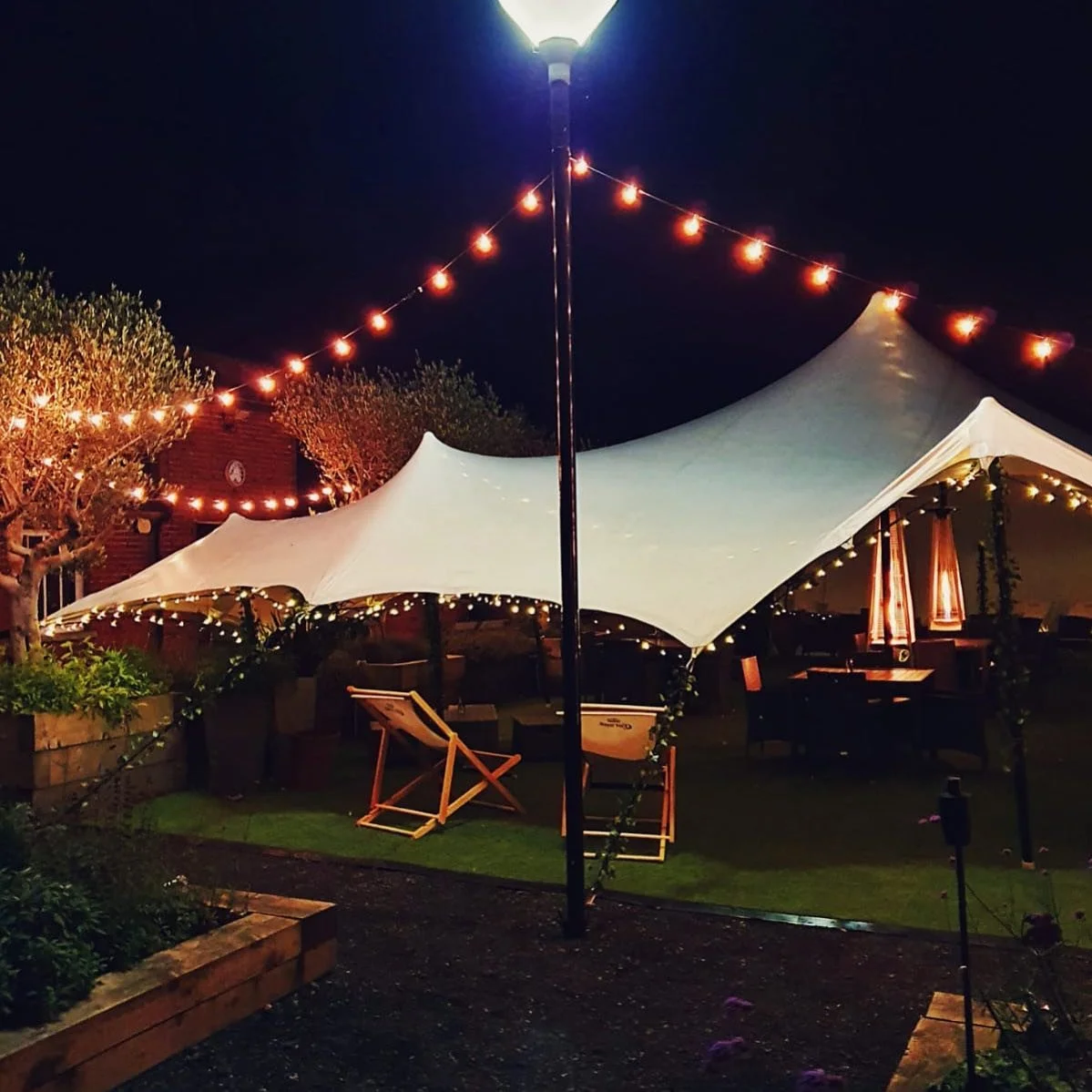 Night scene of outdoor area with a white tent, string lights, and a lamppost illuminating the space. There are tables, chairs, and a covered patio area.