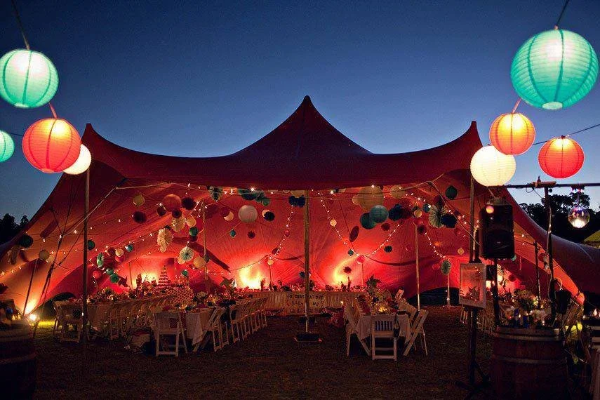 A large red stretch tent decorated with colourful paper lanterns and string lights, set up outdoors during the evening with tables and chairs inside for a festive event.