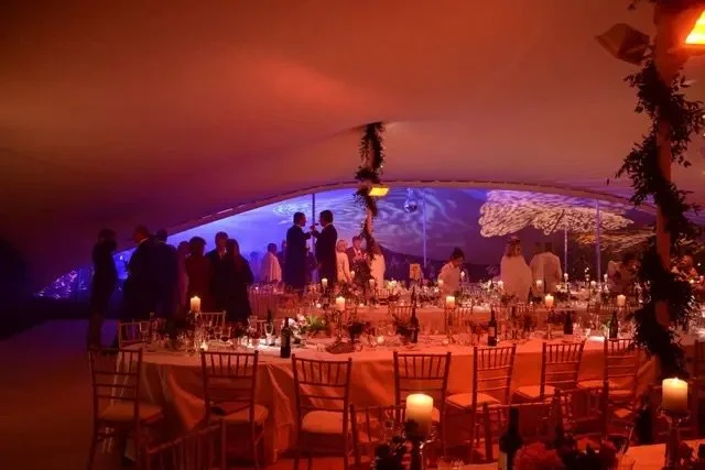 Indoor event with a long banquet table set with plates, glasses, and candles, under a tent with colorful lighting and a view of a cityscape at night.
