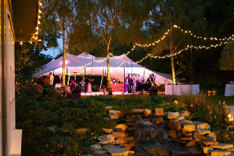 Outdoor evening party under a large white stretch tent with string lights, people socialising near a stage, and a small waterfall feature in the foreground.