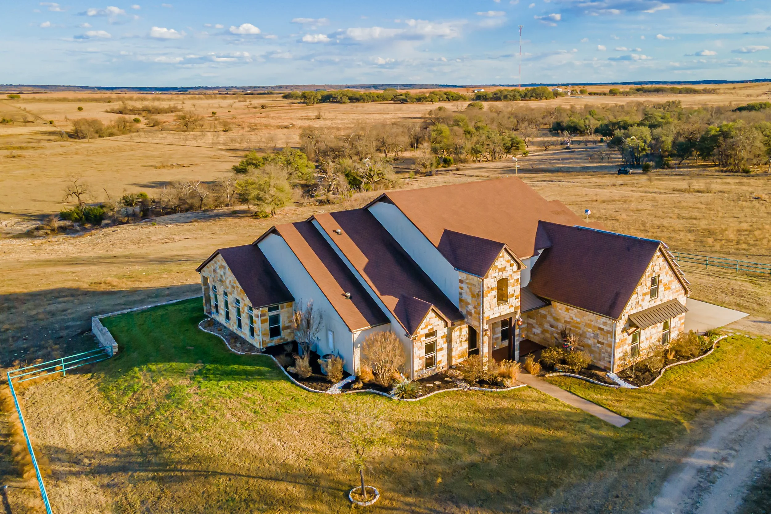 A large house with brick and white siding exterior surrounded by a lawn and garden, located in a rural landscape with open fields and a few trees, under a partly cloudy sky.