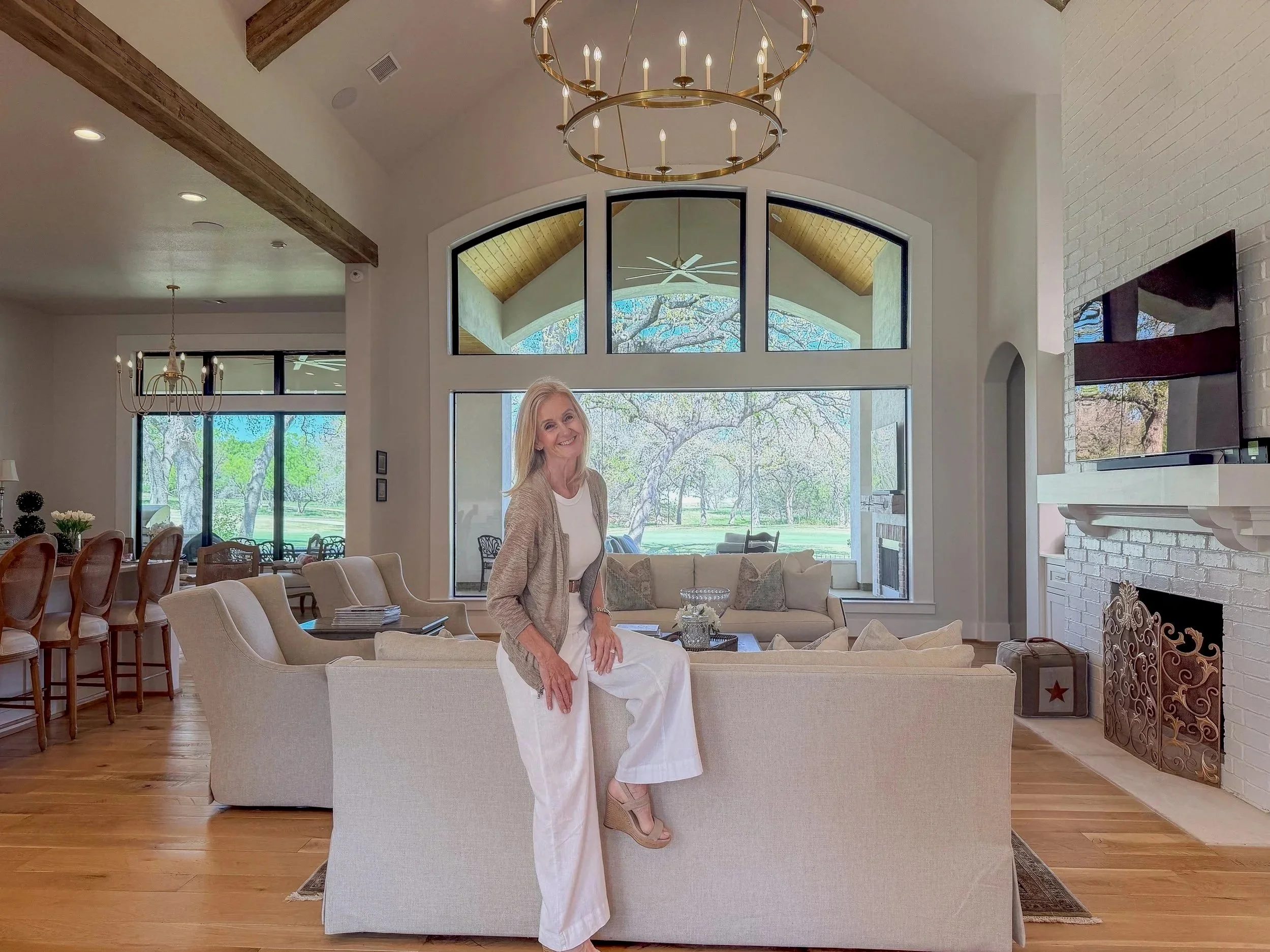 A woman with blonde hair smiling and sitting on the back of a sofa in a bright living room with large windows showing a view of trees and a lawn outside.