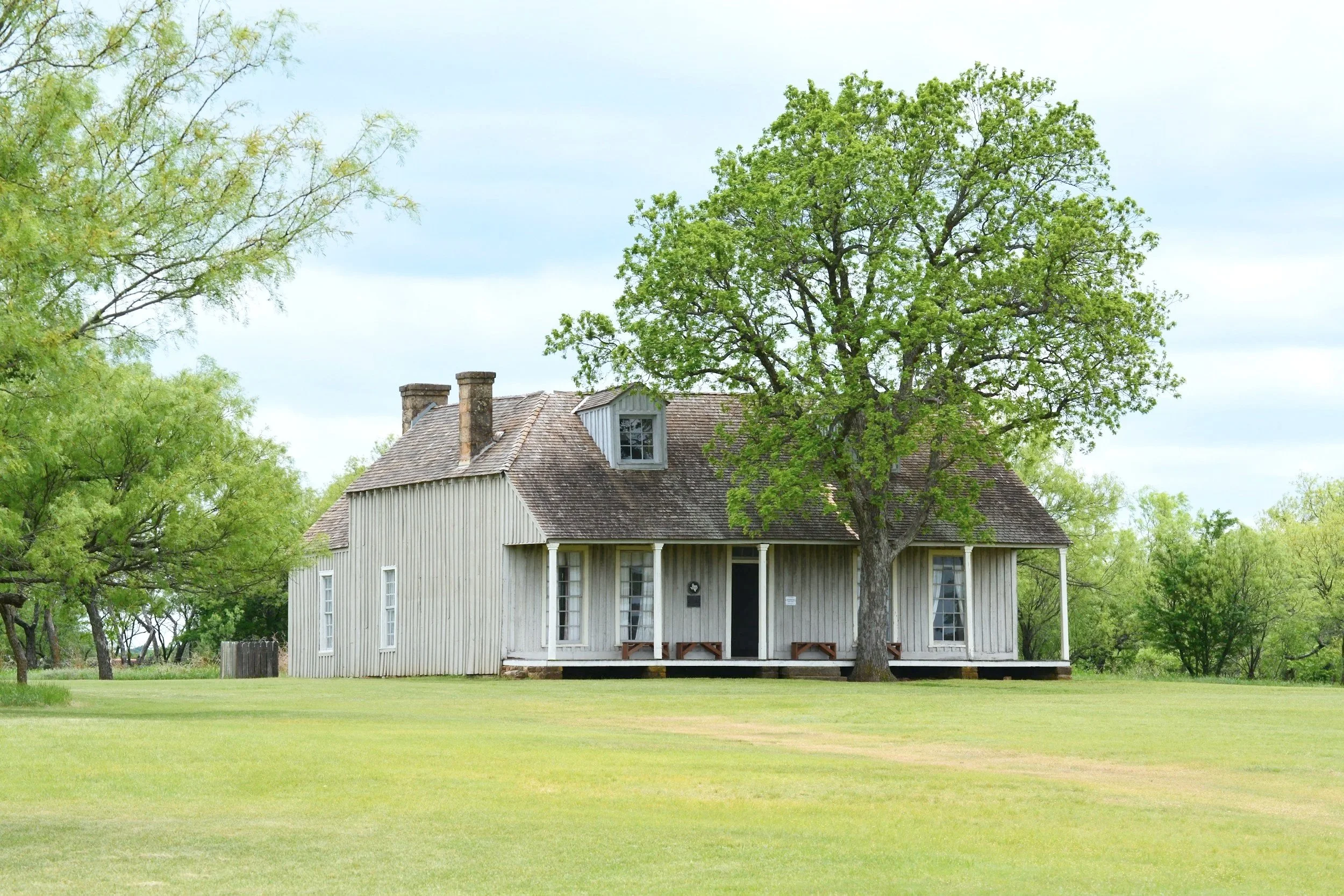 A white wooden house with a porch, surrounded by green trees and a well-maintained grassy lawn.