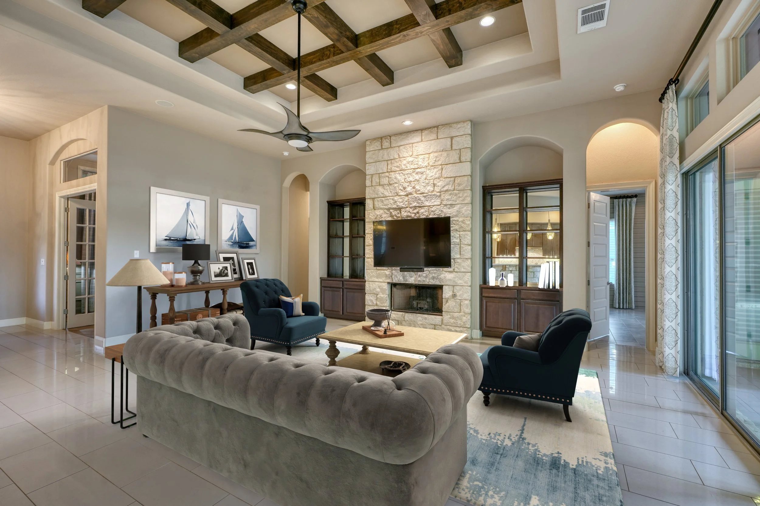 Living room with beige tiled floor, a light gray tufted sofa, two navy blue armchairs, a wooden coffee table, and a stone fireplace with a mounted television. There are built-in wooden cabinets on either side of the fireplace, a ceiling fan with blades, and a coffered ceiling with exposed wooden beams. Large sliding glass doors with patterned curtains open to an outdoor area.