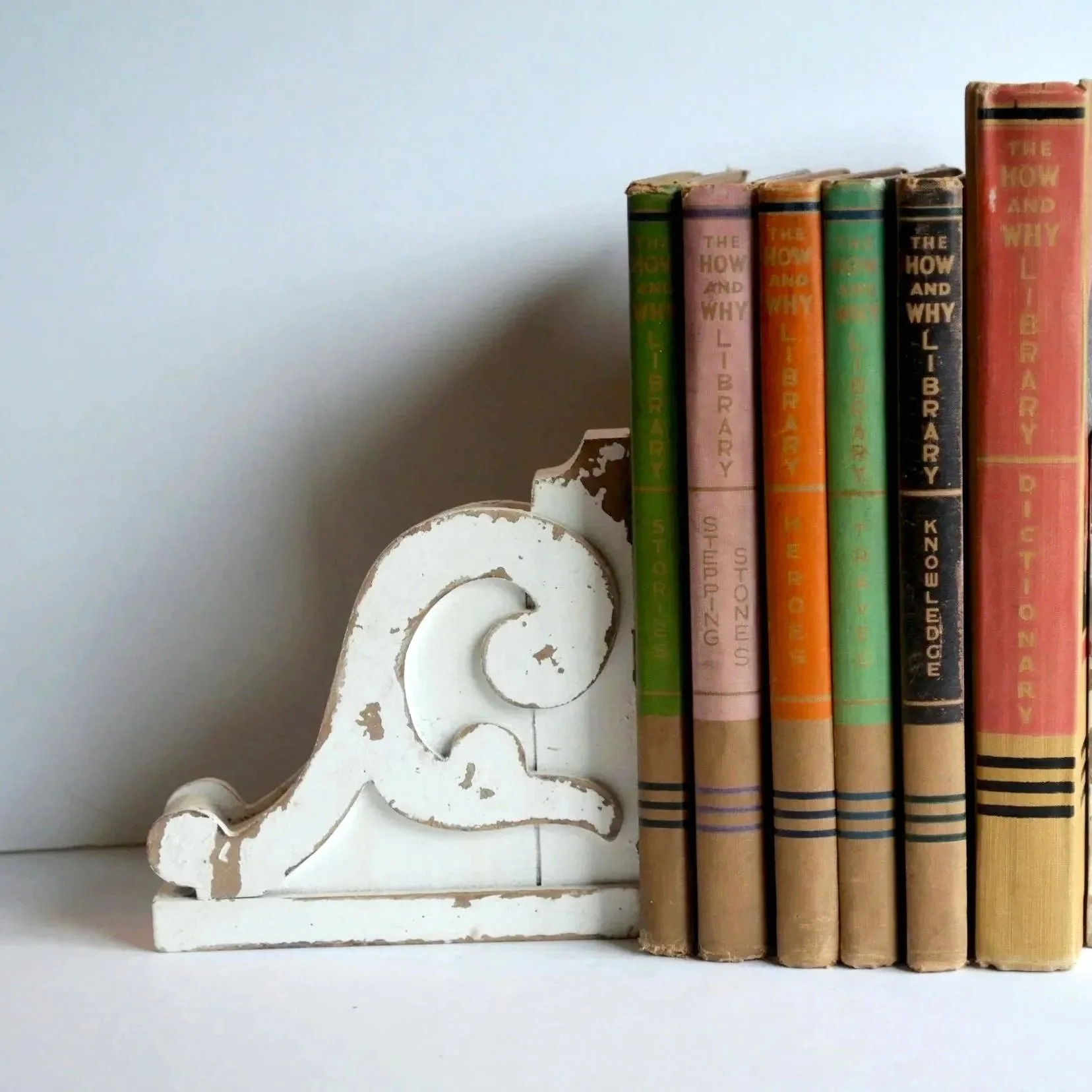 A white, distressed decorative piece in the shape of a lamp next to a row of vintage books with colorful spines on a white shelf.