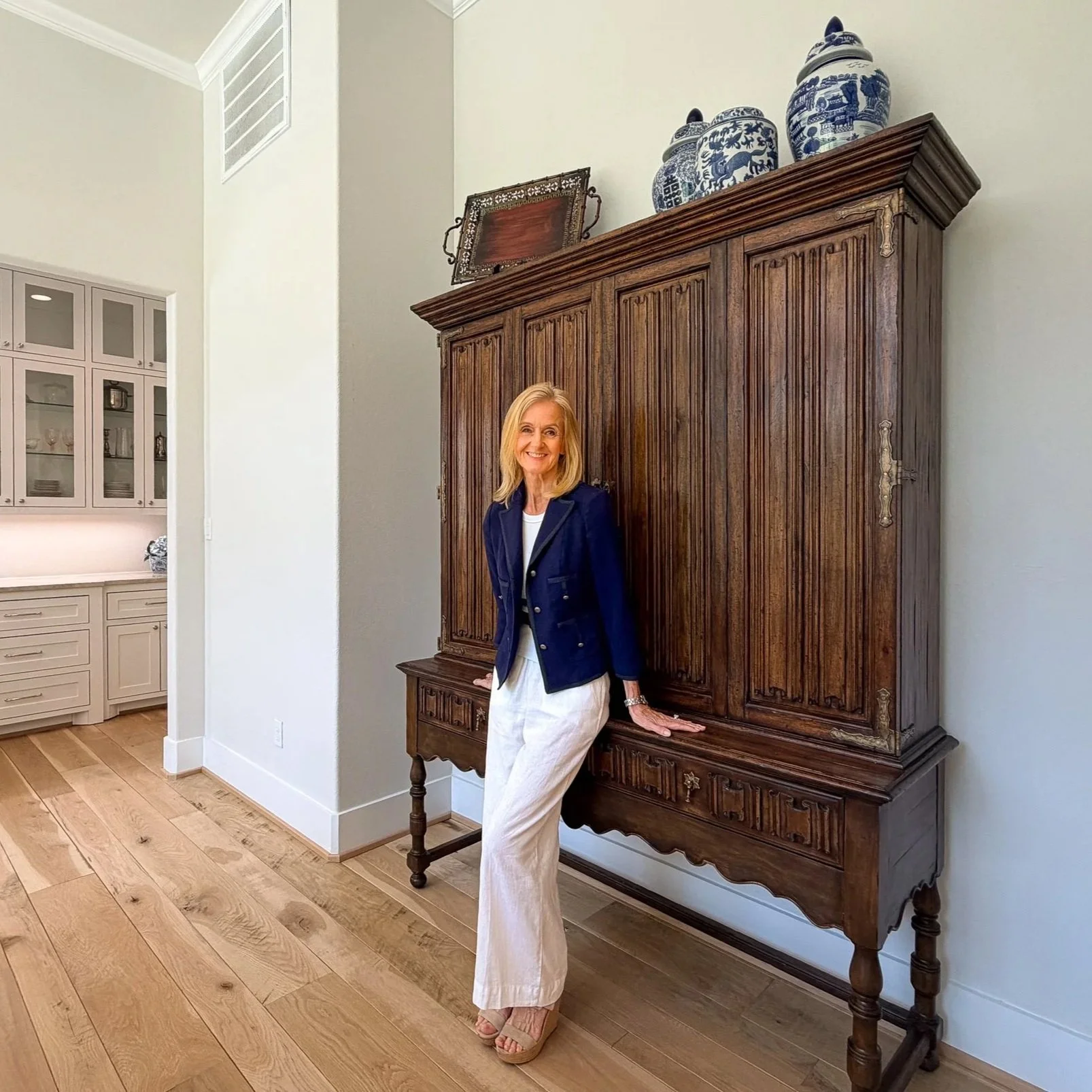 A woman with blonde hair wearing a navy blazer, white pants, and beige shoes, standing and leaning against a large antique wooden cabinet with blue and white porcelain jars on top, in a bright room with light-colored walls and wooden flooring.