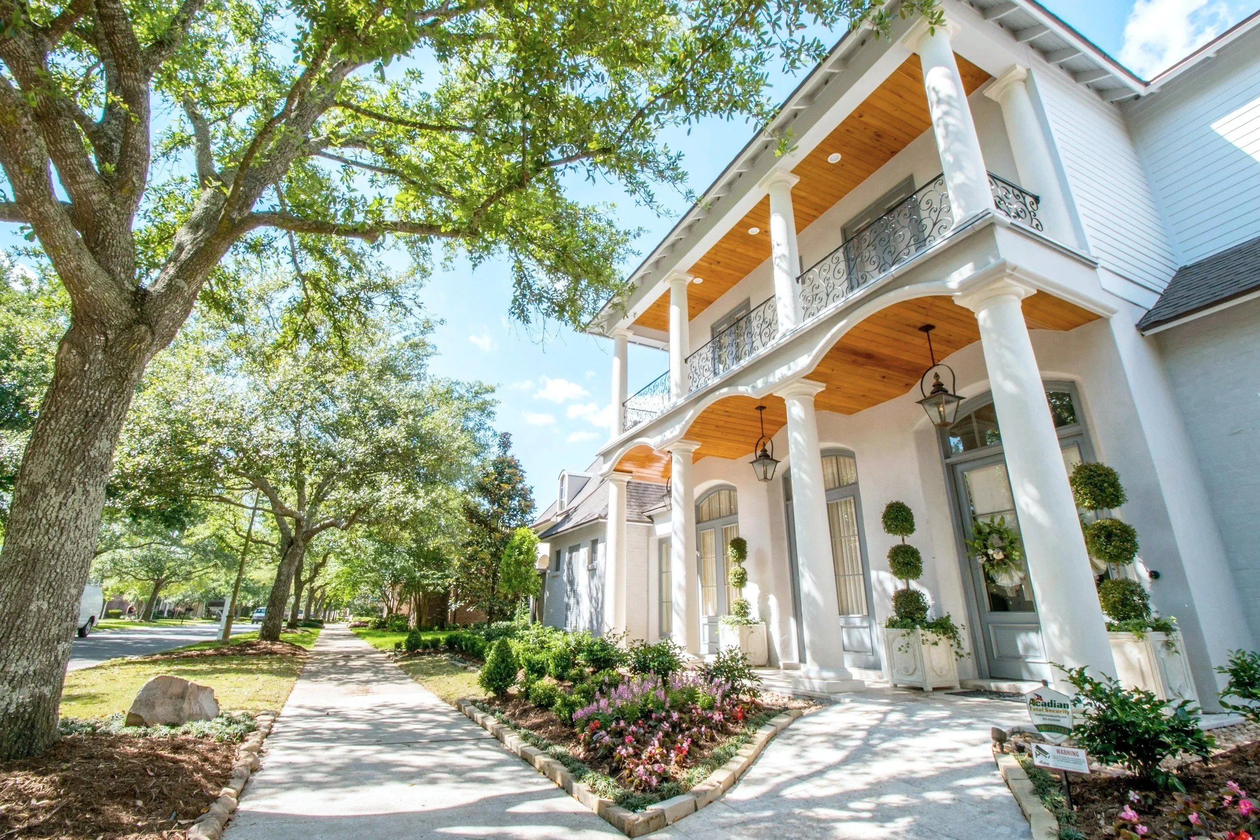 A white two-story house with a front porch and balcony, surrounded by trees and a landscaped garden with flowering bushes, on a sunny day.