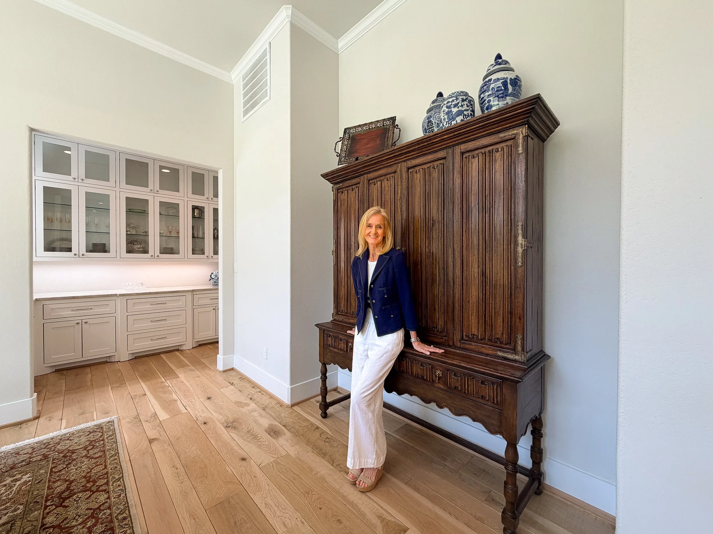 A smiling woman leaning against a large antique wooden cabinet in a room with white walls and hardwood floors.