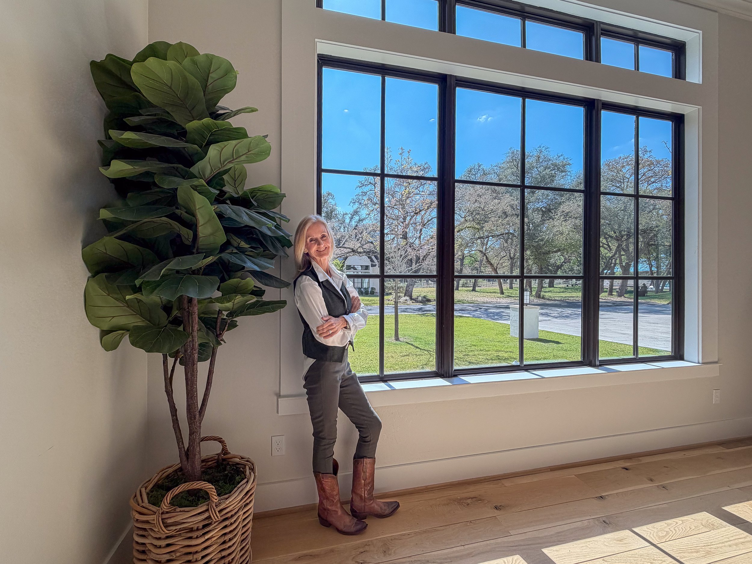 A woman standing inside near a large window with a view of trees and a street, next to a tall potted plant with broad leaves.