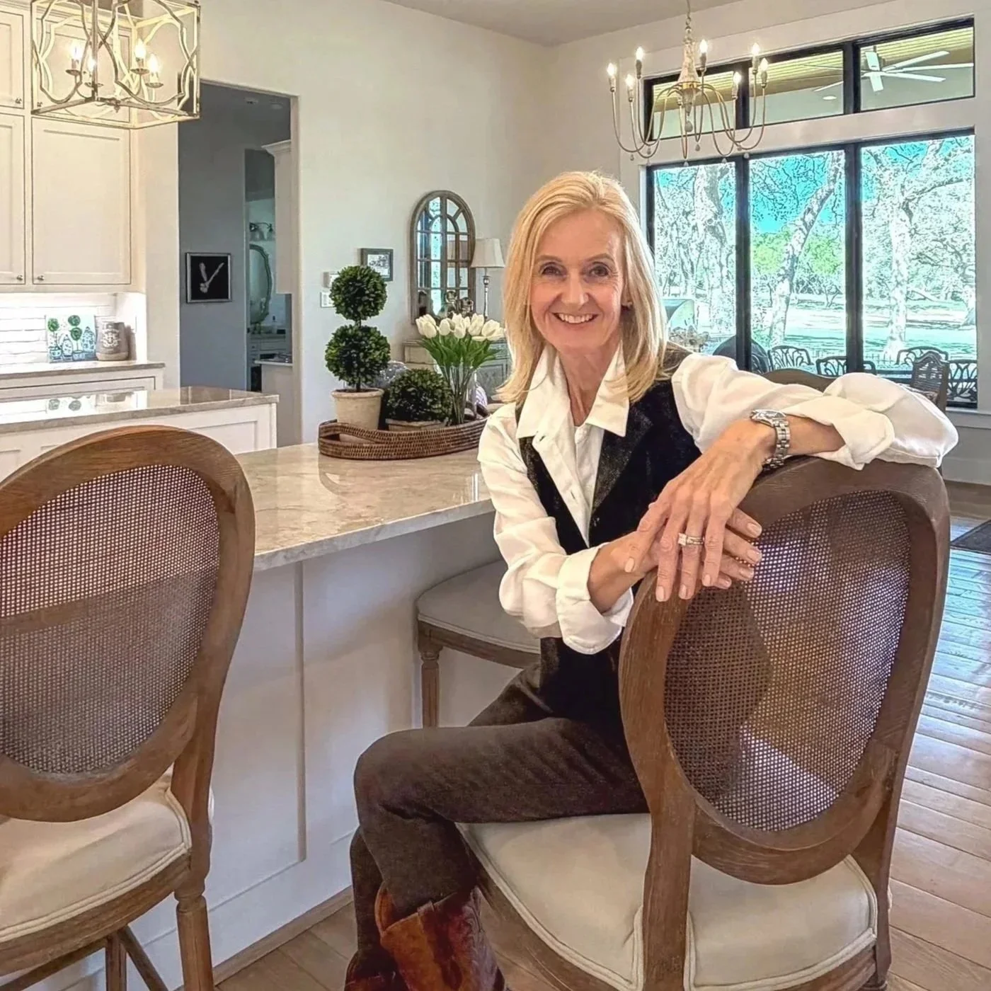 A smiling middle-aged woman sitting on a chair in a bright kitchen with large windows showing trees outside. She has blonde hair, is wearing a white blouse and a dark vest, with her left arm resting on the back of the chair, showing a ring and watch.