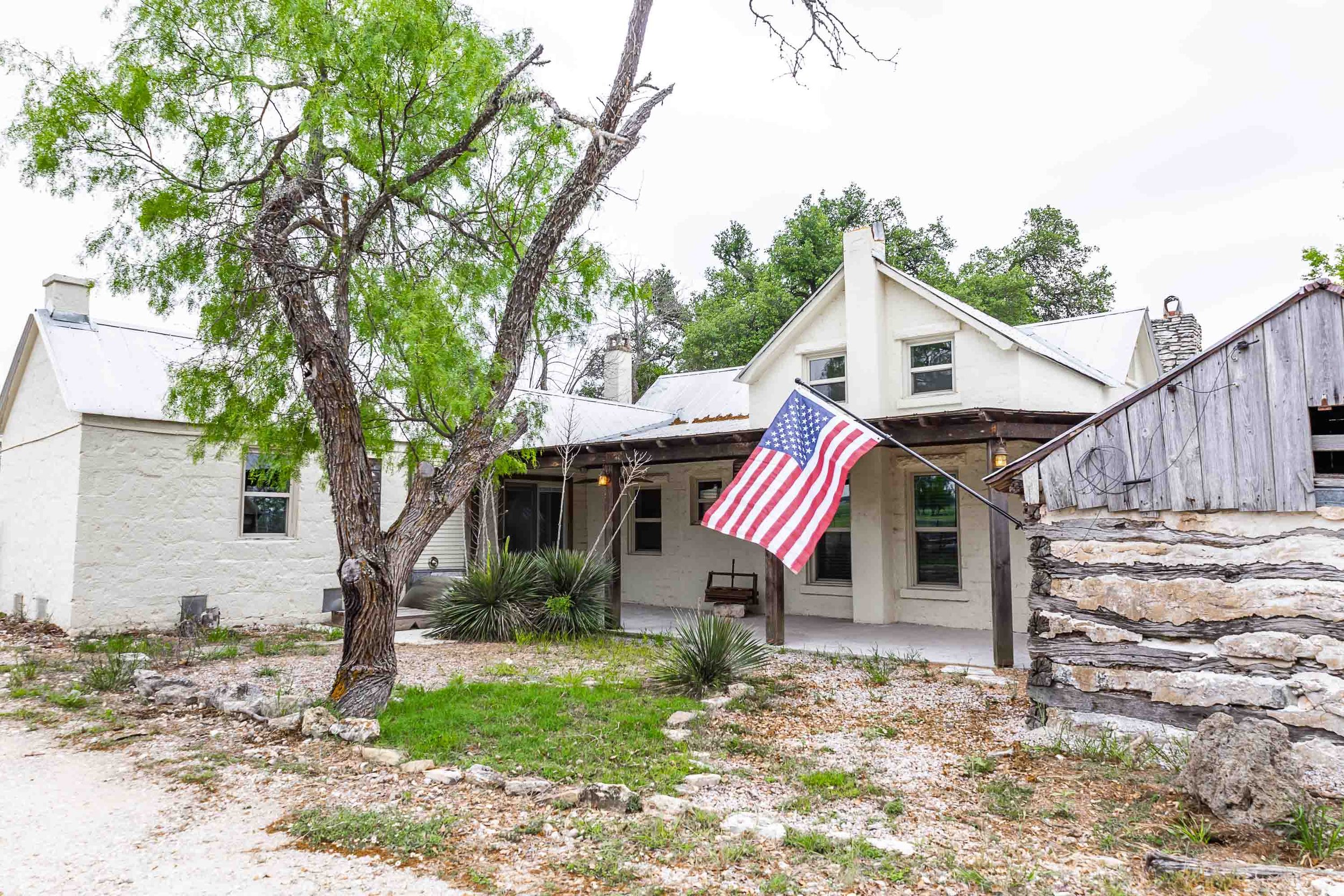 Front view of a house with a large tree in the yard. An American flag is hanging from a pole attached to the house's porch. The house has a white exterior with multiple windows and a porch area. The yard has some grass, bushes, and rocks. The sky is overcast.