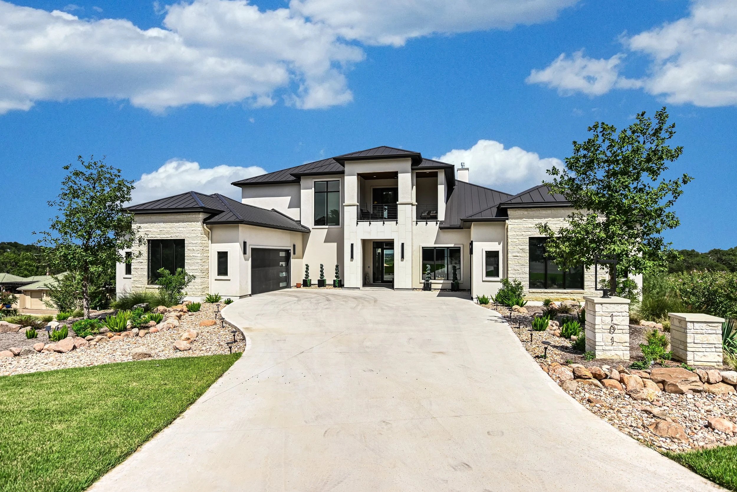 Modern two-story house with a black metal roof, white walls, large windows, and a circular driveway, surrounded by landscaped yard with trees and rocks, under a partly cloudy blue sky.