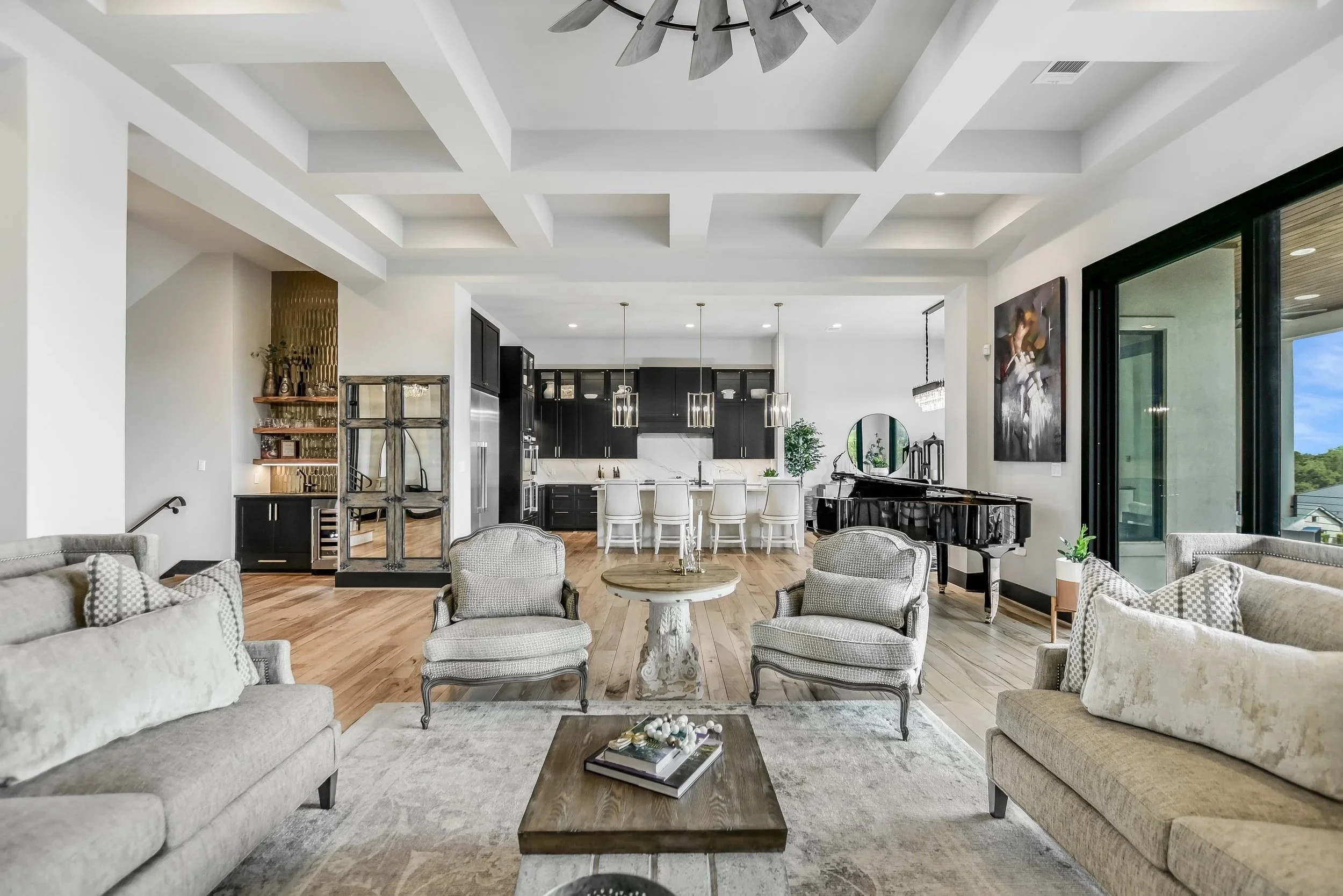 Living room and kitchen with white walls, dark cabinetry, a black grand piano, and large windows showing an outdoor view.