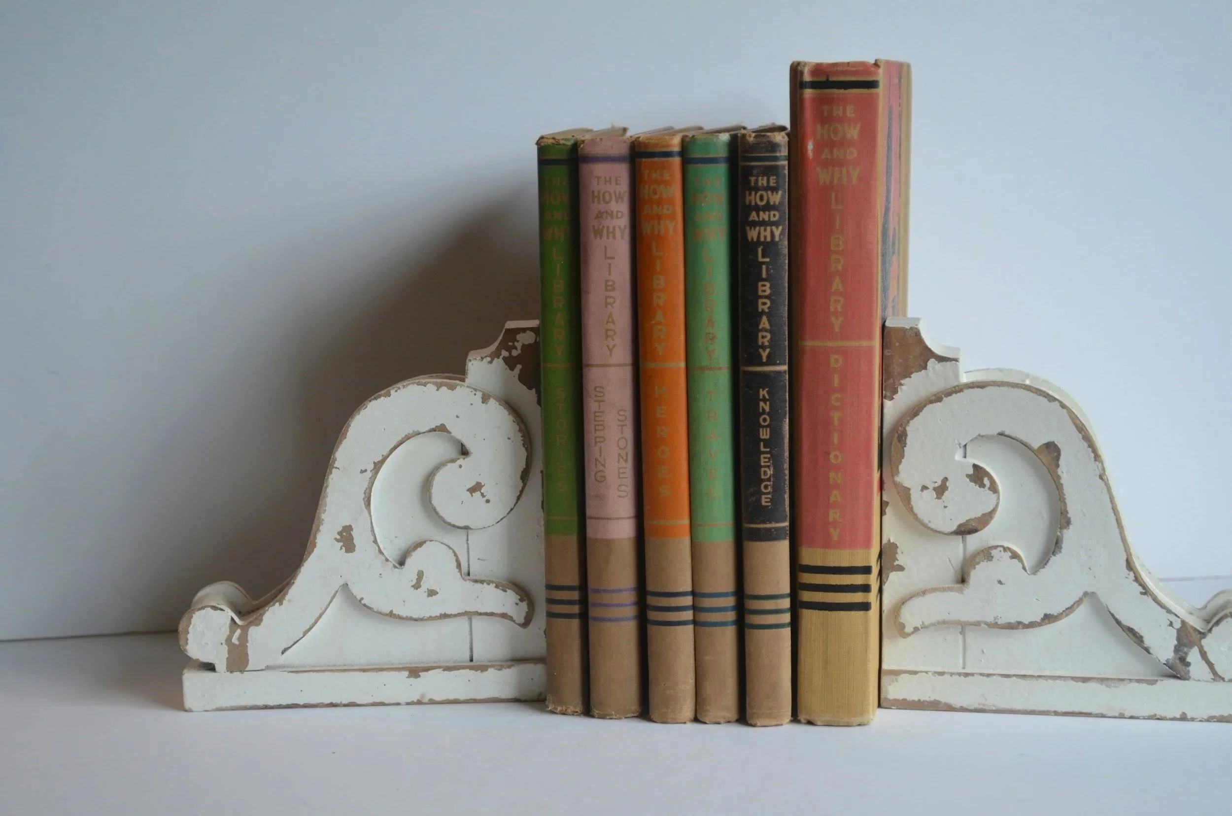 Vintage white wooden bookshelf with ornate scrollwork design, holding a row of old, worn books with colorful spines including titles such as 'The How and Why Library' and 'Dictionary'.