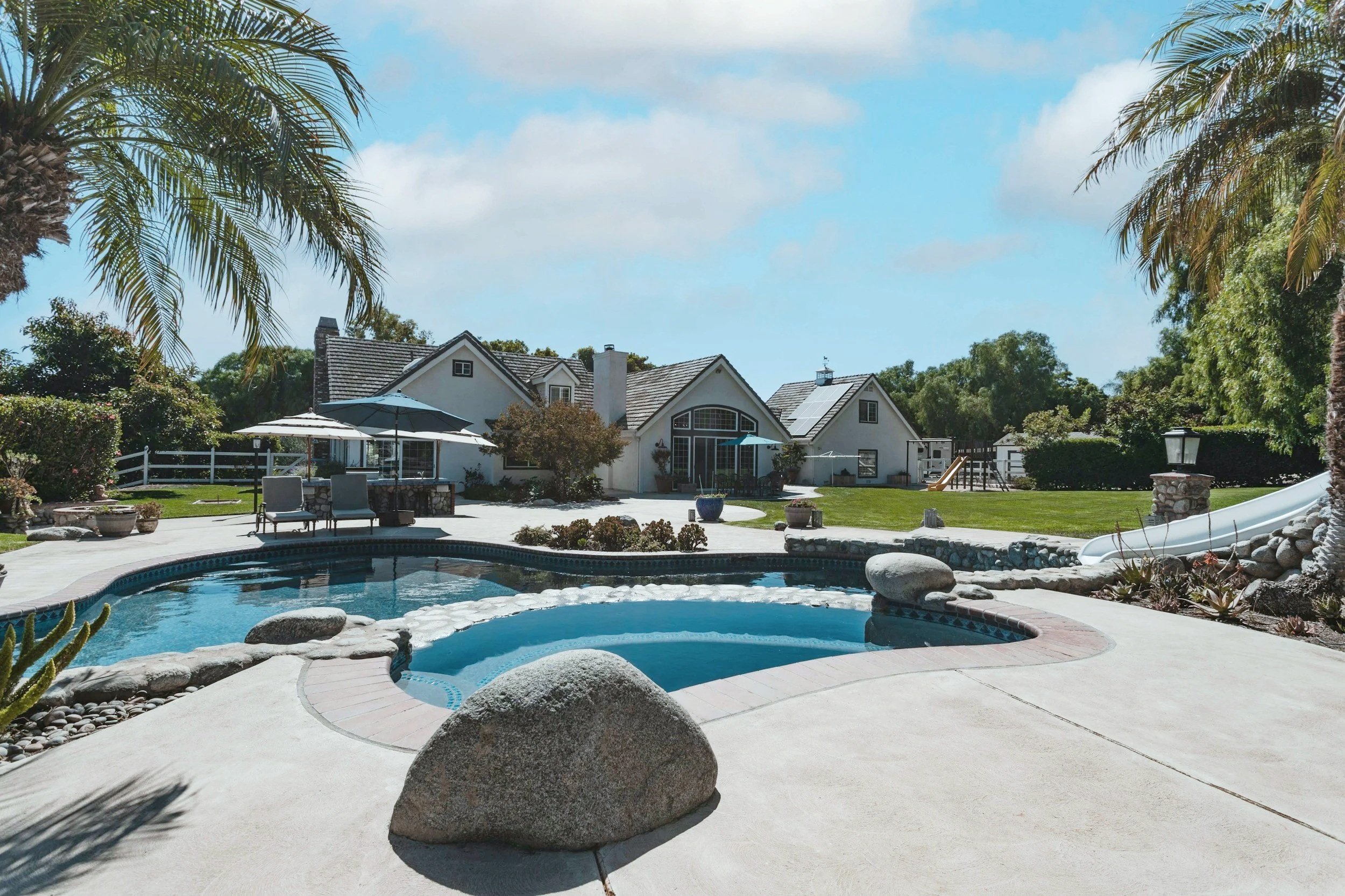 A backyard with a swimming pool, umbrella-shaded seating area, well-kept lawn, and a large white house with multiple gabled roofs, surrounded by lush greenery and palm trees.