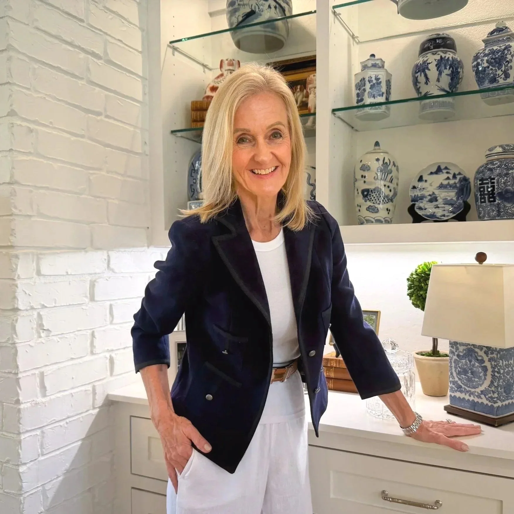 A smiling woman with blonde hair, wearing a navy blazer and white pants, leaning on a white desk in front of a display of blue and white porcelain vases and jars on glass shelves.