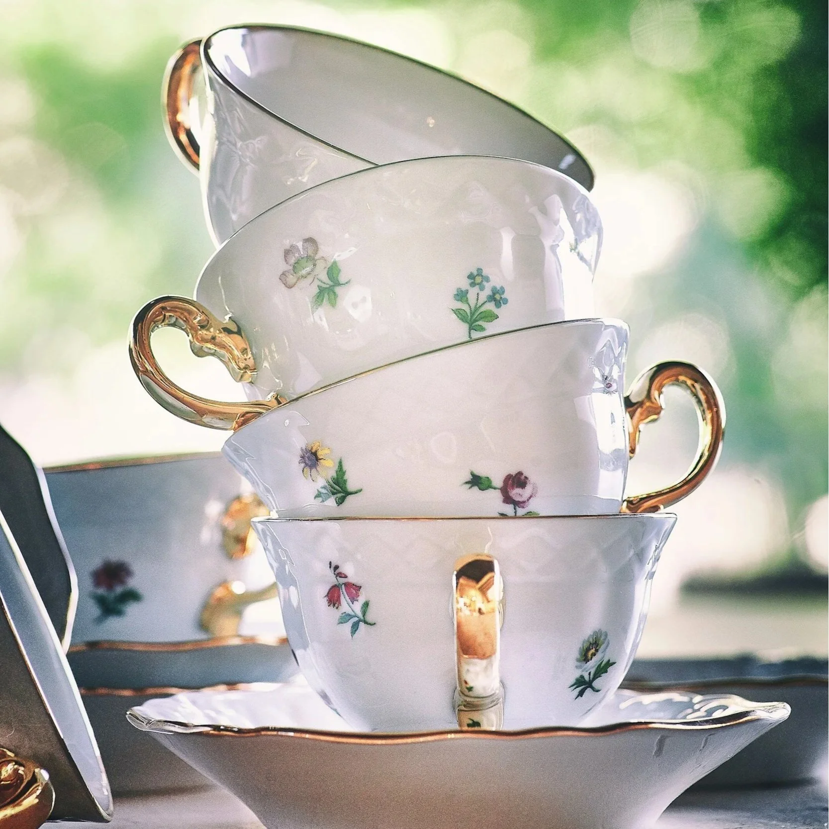 Stacked fine china teacups with floral patterns and gold handles on a matching saucer, with a blurred green background.