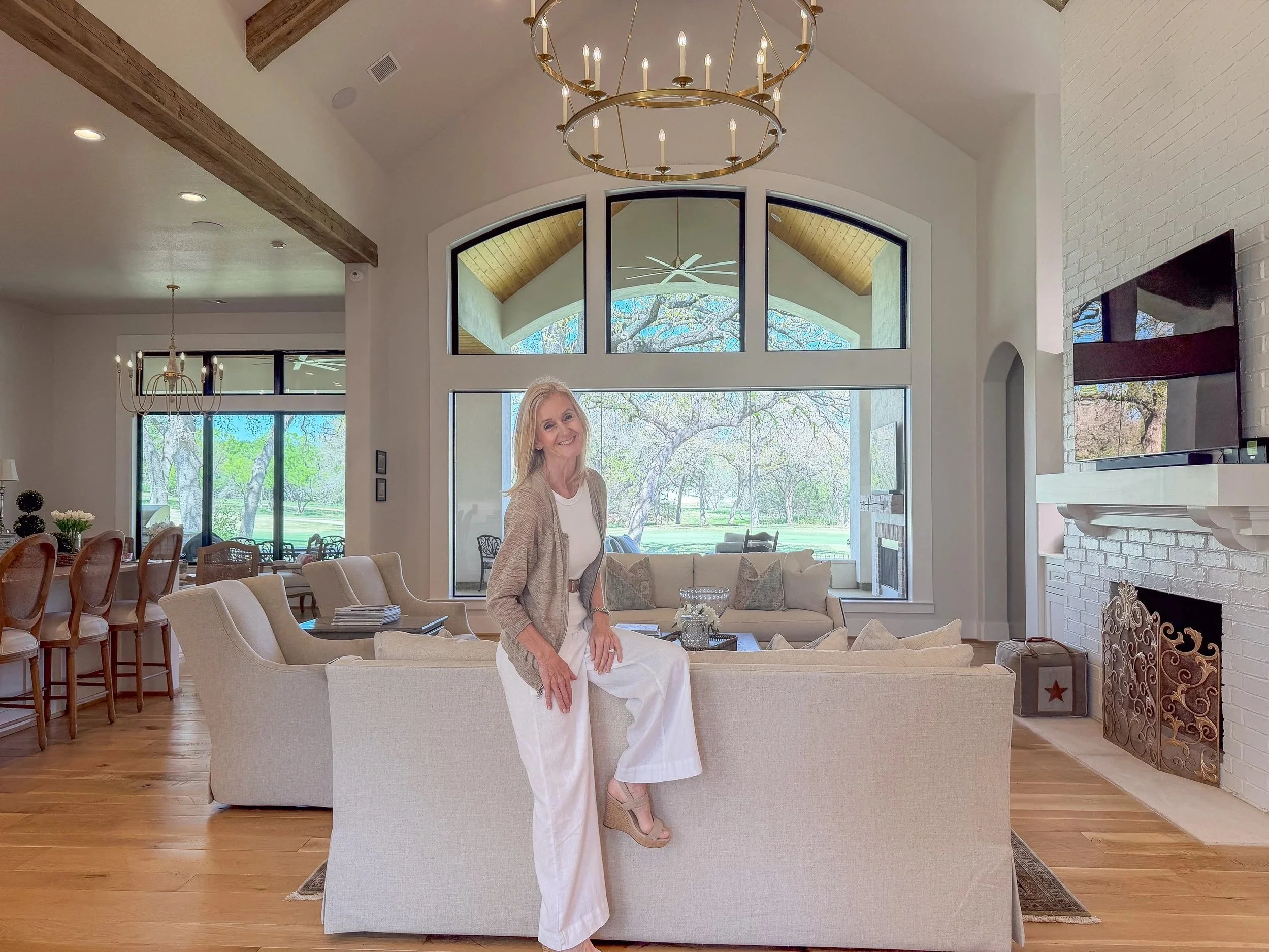 A woman sitting on the back of a beige sofa in a spacious, well-lit living room with large windows showing trees and a lawn outside. The room has white walls, wooden floors, a black wall-mounted TV, a fireplace, and a dining area with chairs in the background.