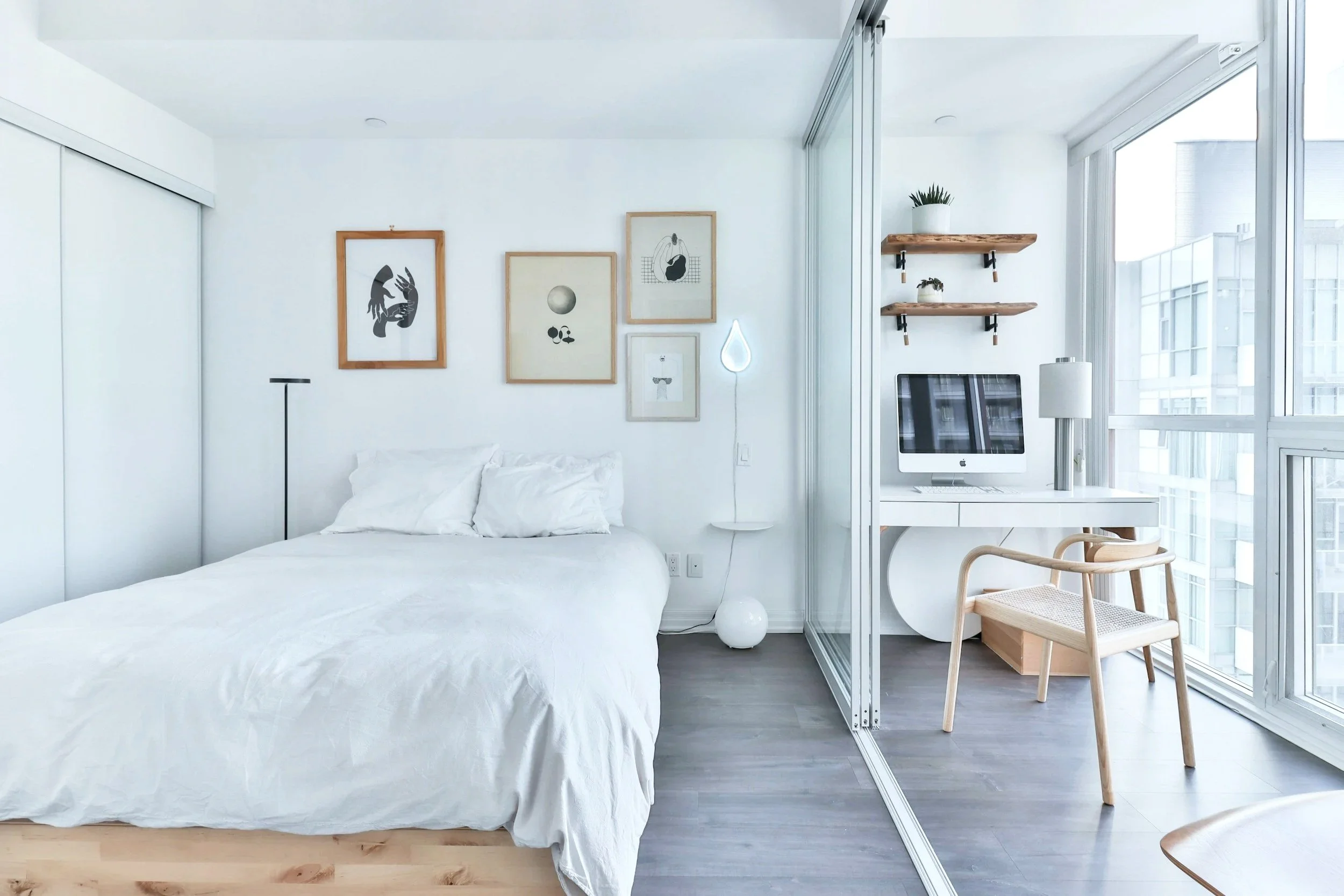 A modern, minimalist bedroom with a white bed, artwork on the wall, a small white side table, and a workspace with a computer and wooden chair near large glass windows.