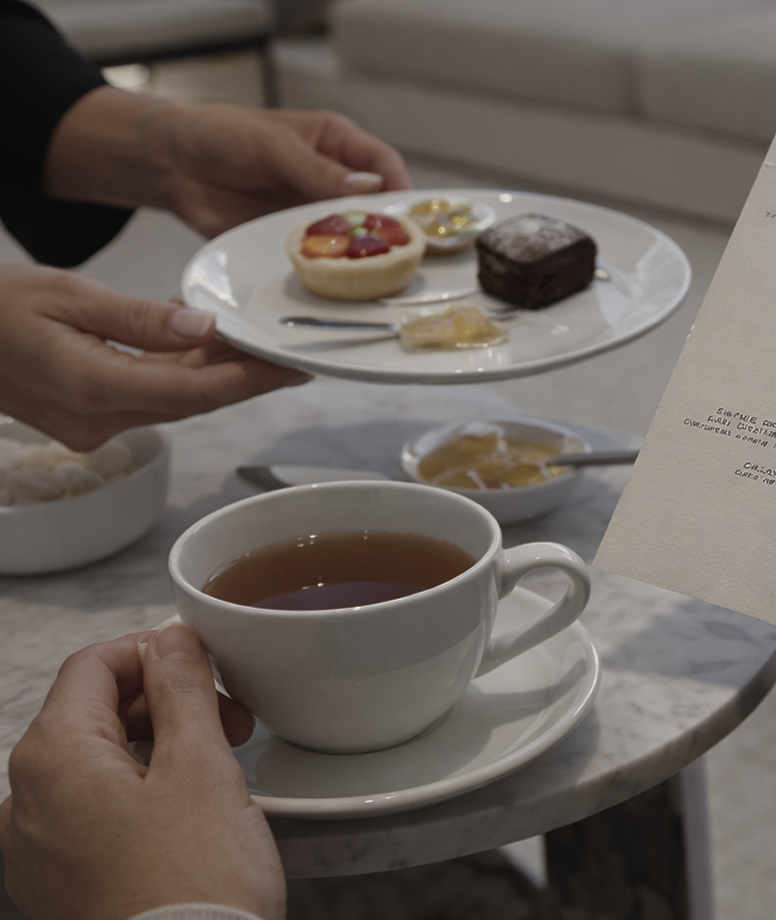 Pessoa segurando uma xícara de chá, com um prato de sobremesas ao fundo, incluindo um brownie e sobremesas com frutas e chantilly, em um ambiente de café ou restaurante.