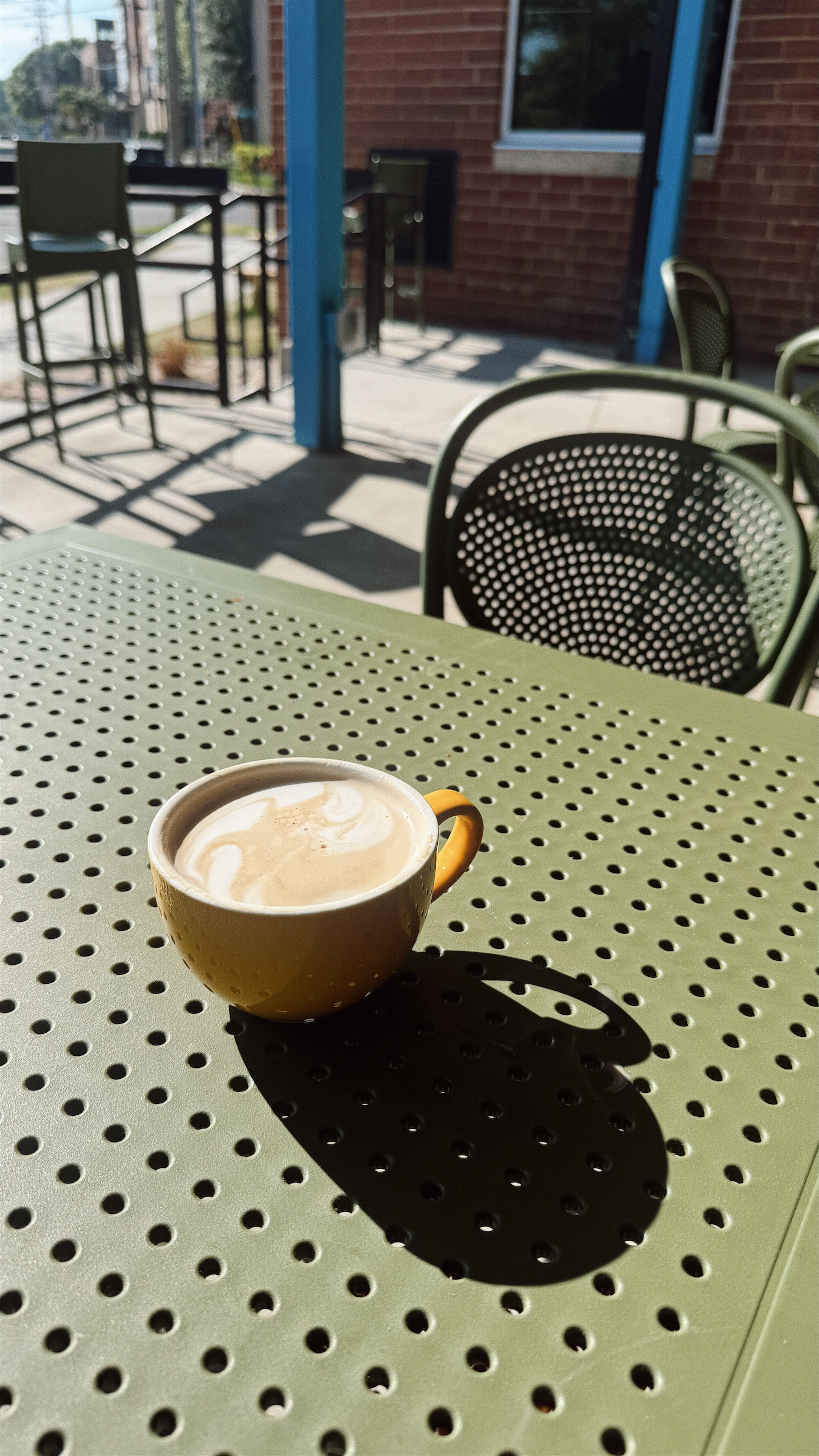 A yellow cup of coffee on a perforated green table outside on a patio, with chairs and a brick building in the background.