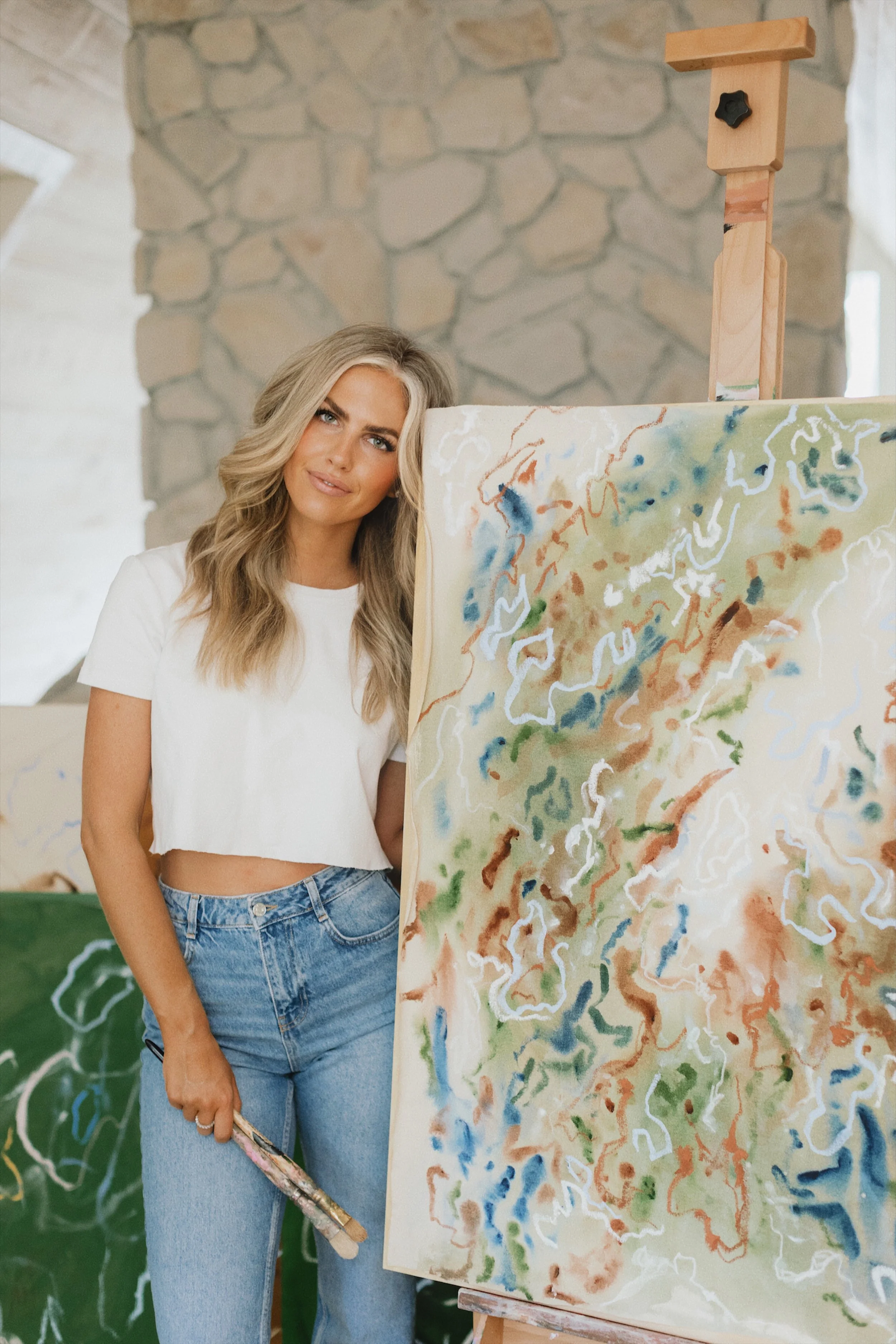Woman with blonde hair in a white crop top and jeans standing next to an abstract painting with colorful, flowing lines on an easel in an art studio.