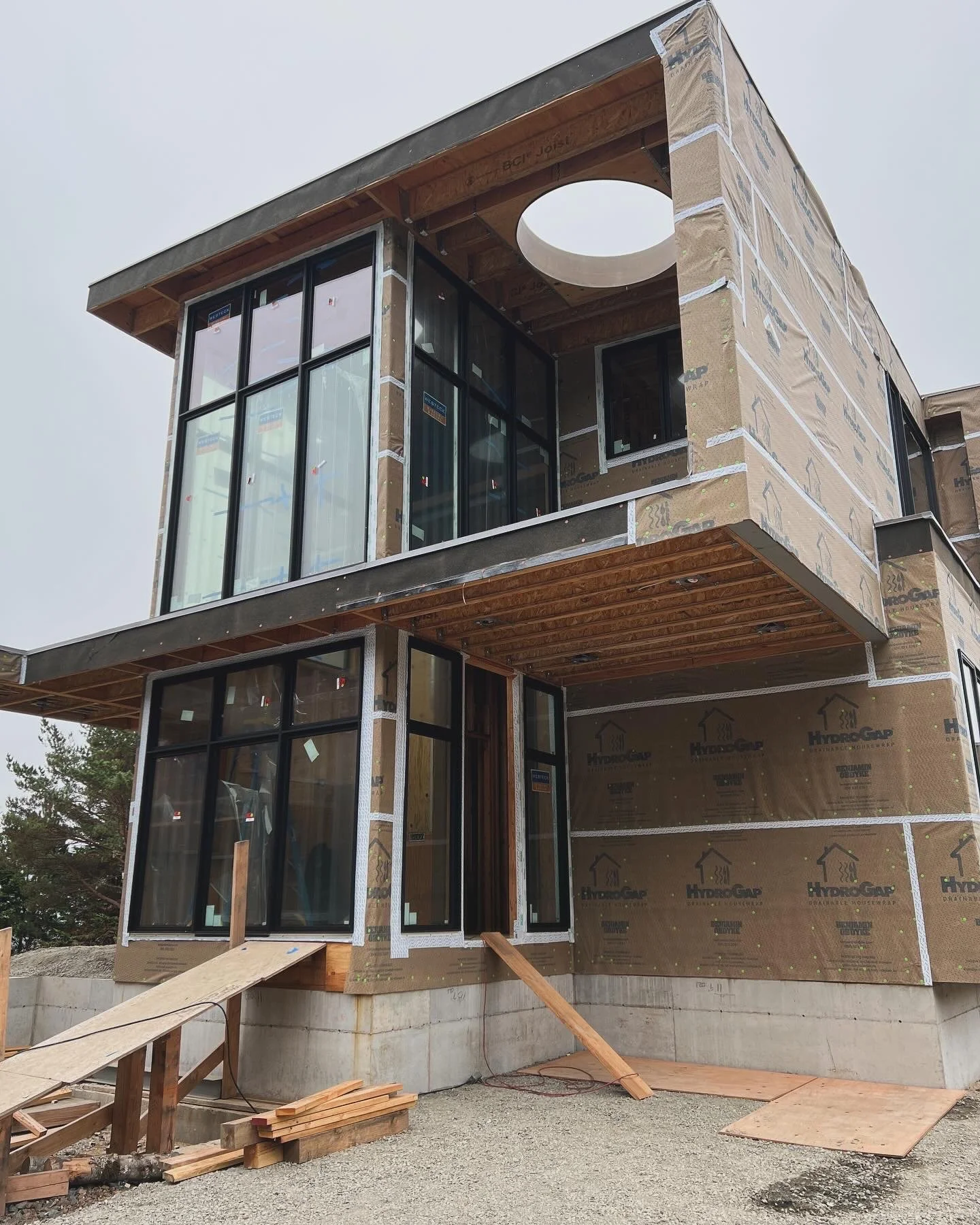 A modern multi-story house under construction with large glass windows, wooden accents, and a circular cutout in the upper section, with construction materials and scaffolding around the building.