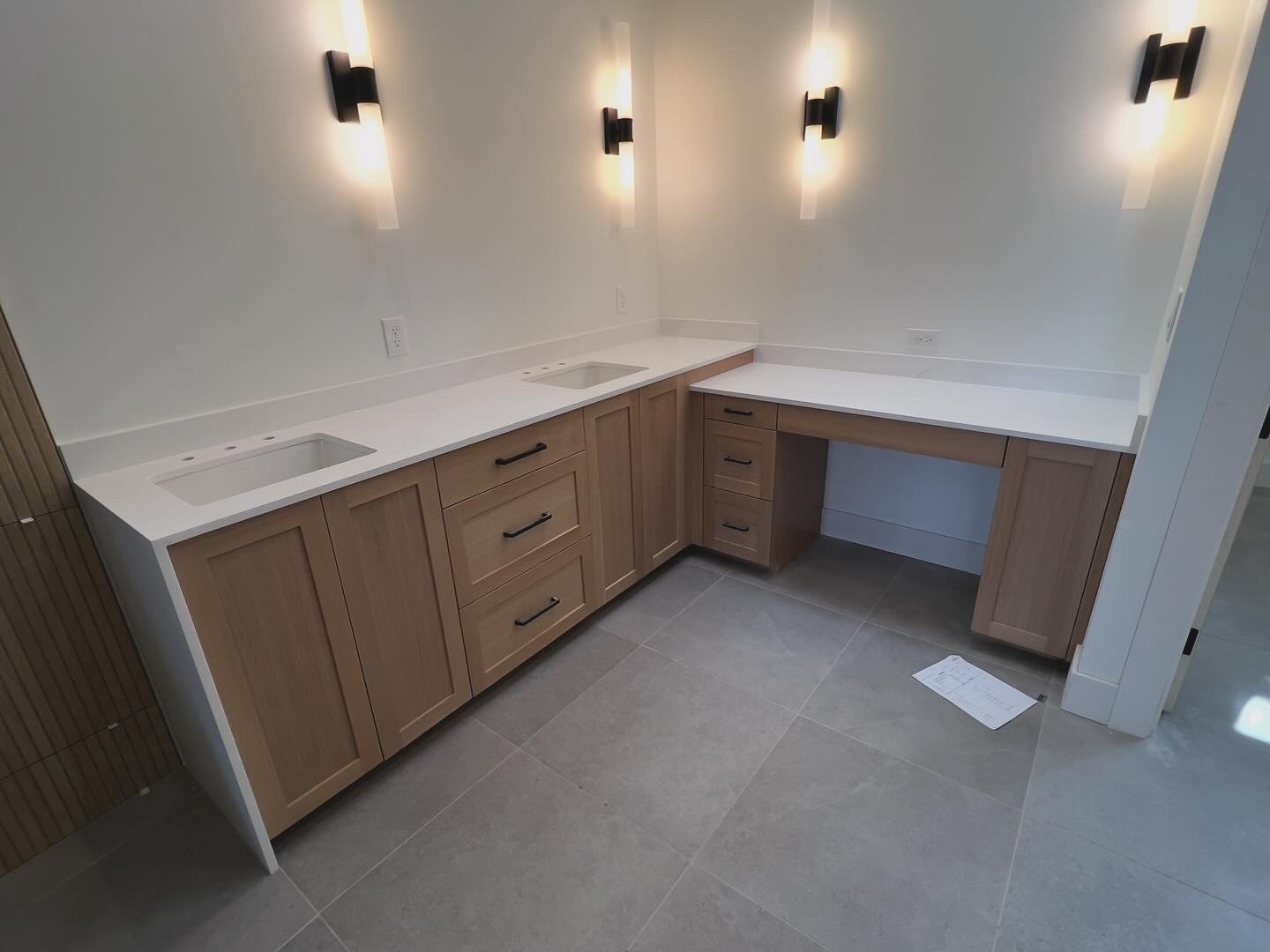 Kitchenette with wooden cabinets, white countertop, and two empty cutouts for sinks, against a white wall with four black wall sconces.