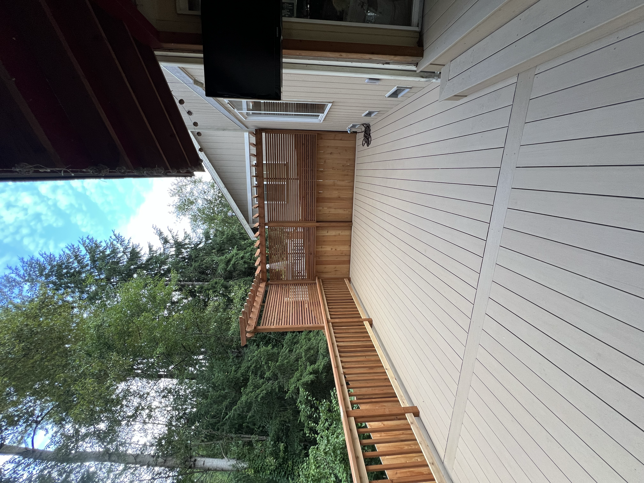 Wooden deck with stairs and railing, surrounded by trees and a house with a sliding glass door, outdoor furniture, and a TV mounted on the house wall.